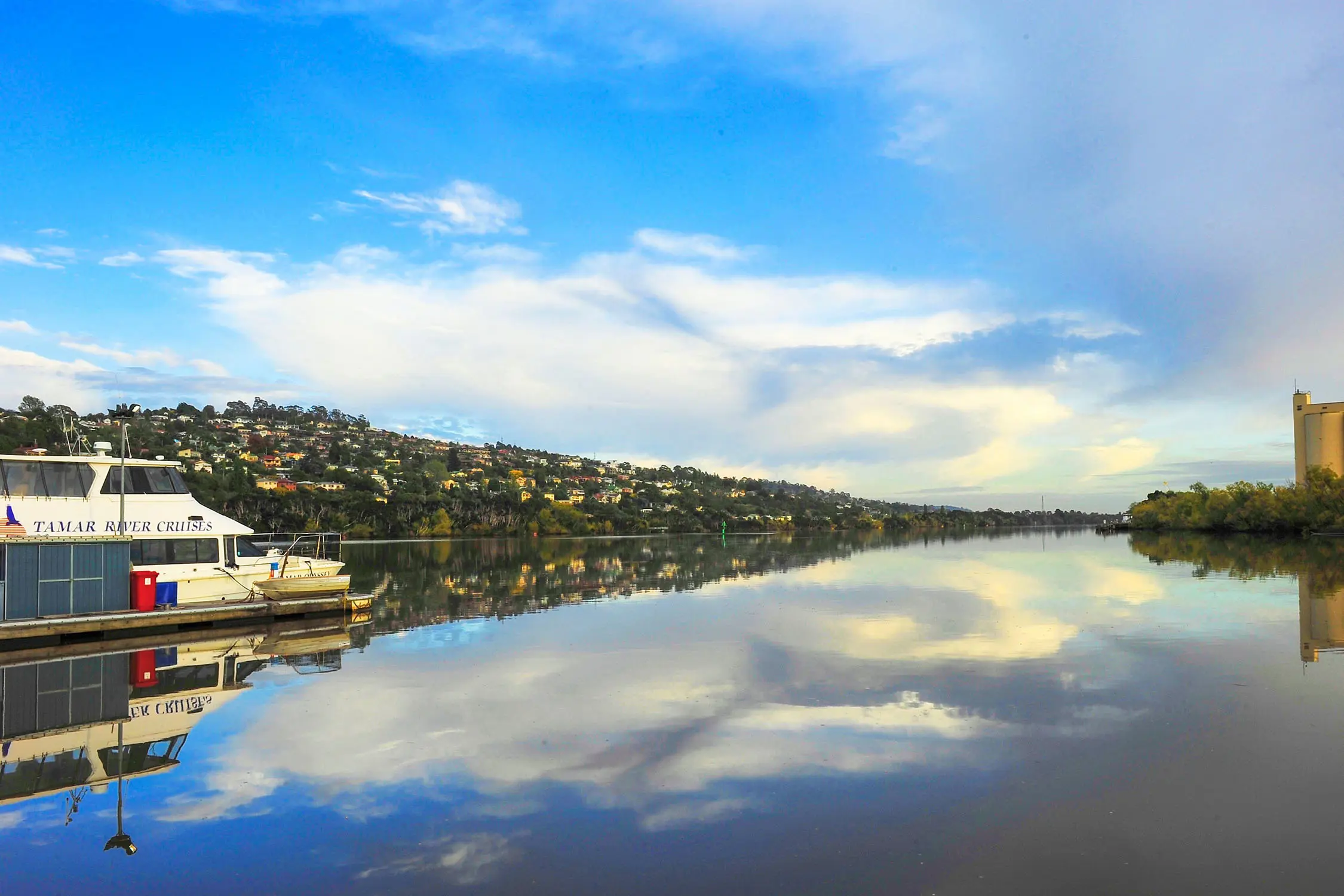 A small river cruise boat in the water, which reflects the blue sky above on its glassy still surface. Houses dot the hillside over on the bank.