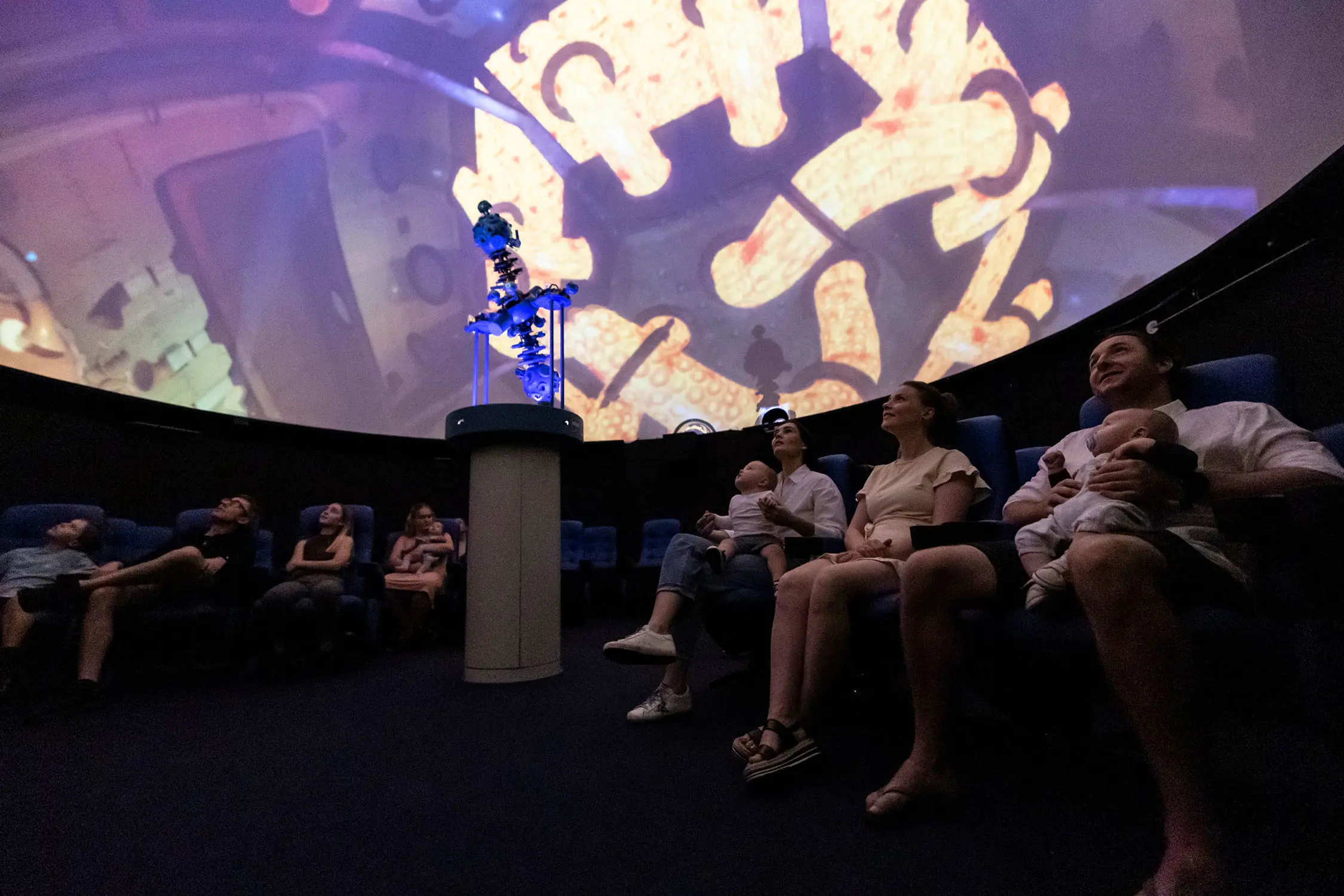 Families sit in a small theatre-like planetarium space, looking up at the images of space projected onto the dome above them.