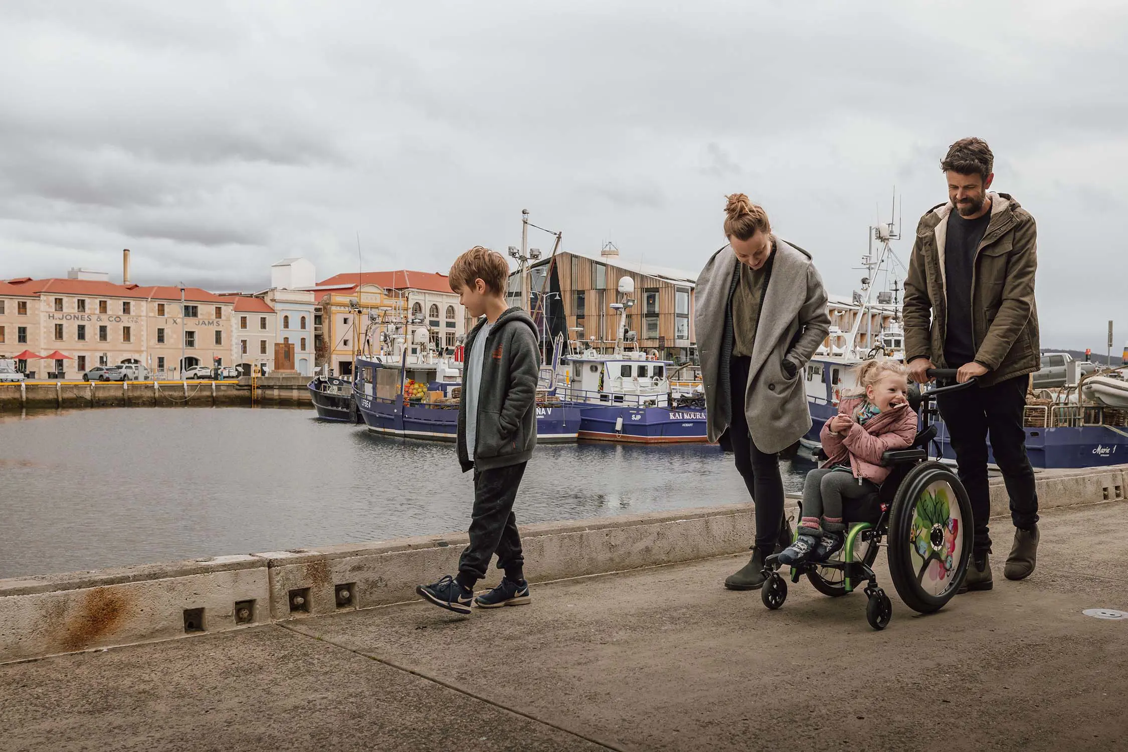 A family of four stroll along Hobart's waterfront, with the little girl in a colourful wheelchair. Historic sandstone buildings and moored boats are in the background.