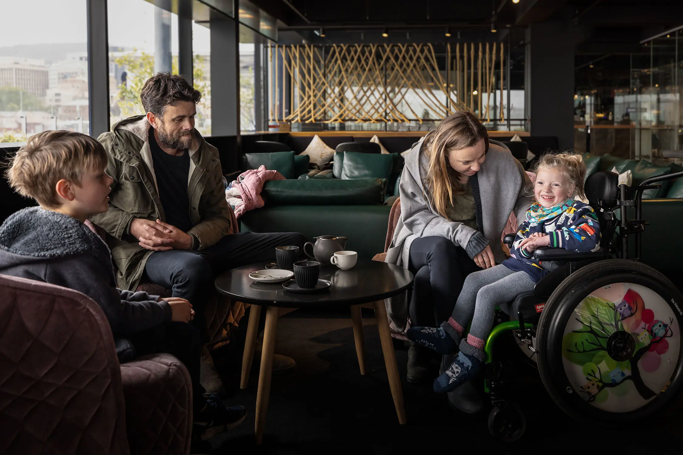 A family of four enjoying tea at the MACq01 hotel. A small girl in a colourful wheelchair smiles.