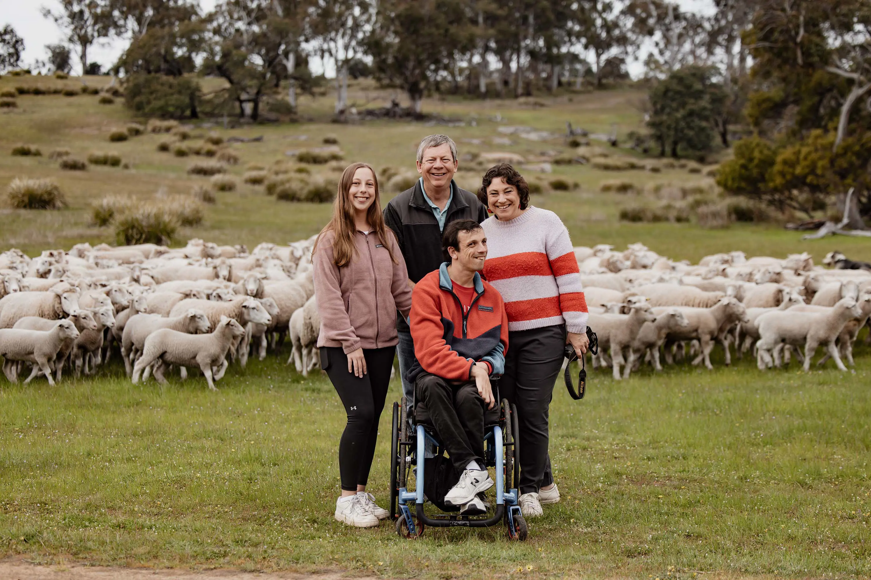 Four people pose for a photo on the grass in front of a herd of sheep. One man sits in a teal blue wheelchair.
