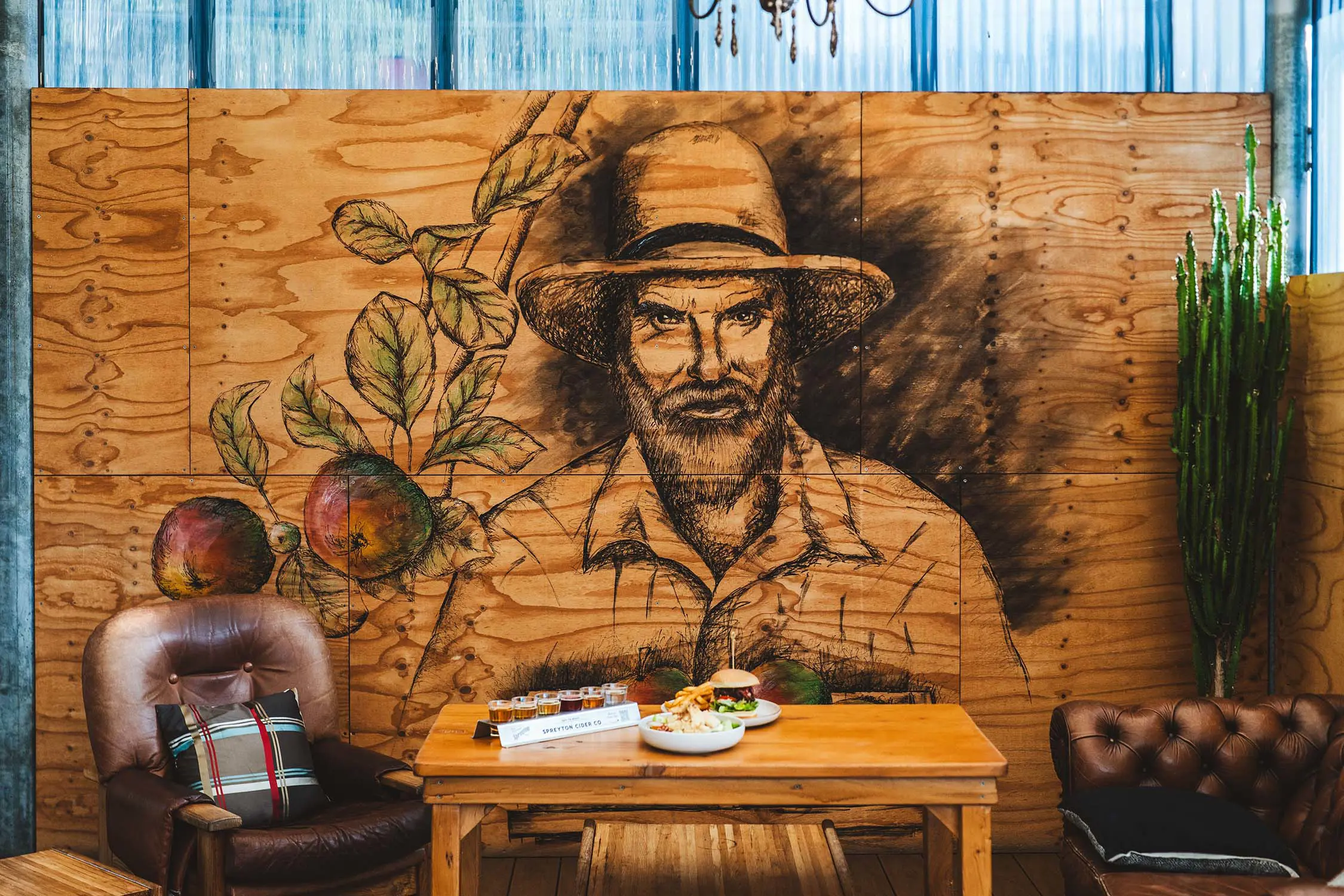 In front of a mural of an apple farmer painted on some wooden boards sits two comfy, worn leather chairs next to a table with plates of burgers and chips.