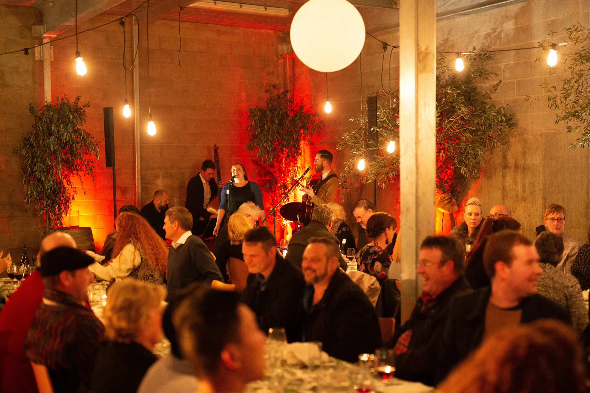 In the corner of a rustic restaurant, a trio of musicians perform. The long tables are filled with people talking and drinking.