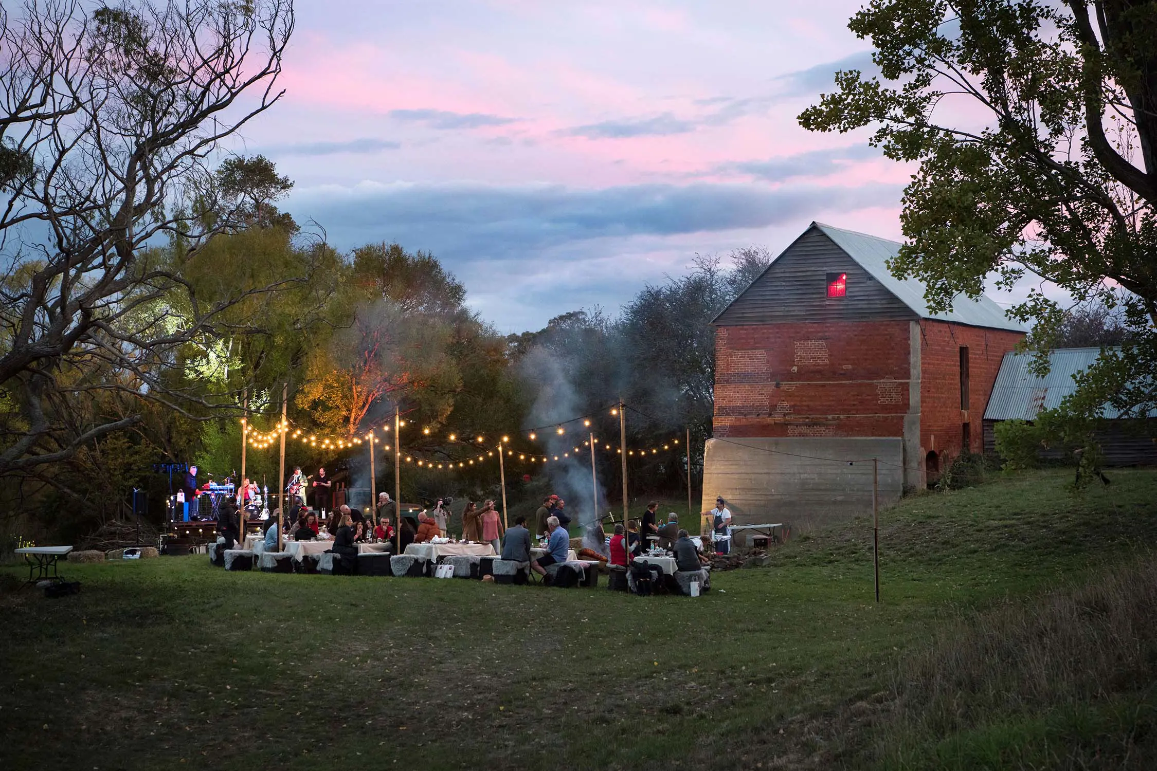 Nestled in a bush clearing next to an old barn-like building, people at rows of long tables under fairy lights watch a live music performance.