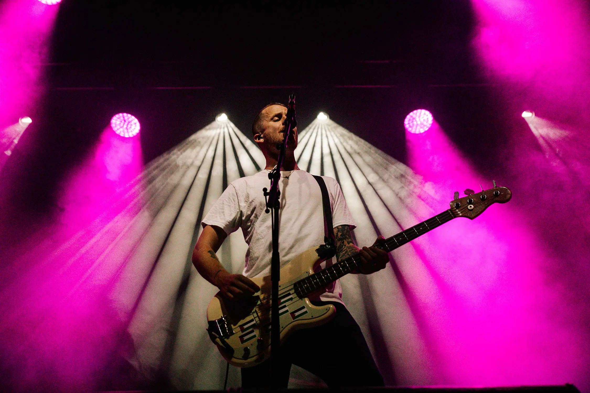 A man in a white tshirt plays an electric guitar and sings into a microphone stand onstage. Behind him, rays of purple and white stage lights catch the stage smoke wafting through the air.