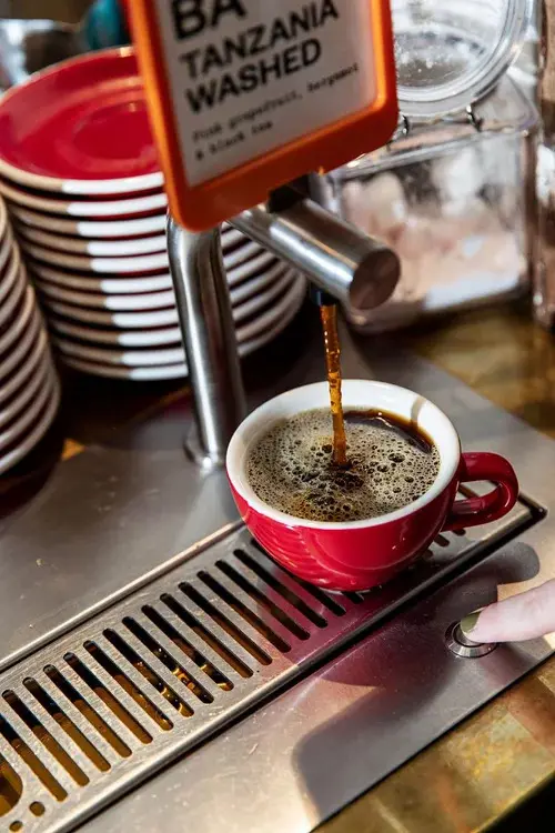 A close up image at a barista's bench of a silver tap dispensing coffee into a red coffee cup.