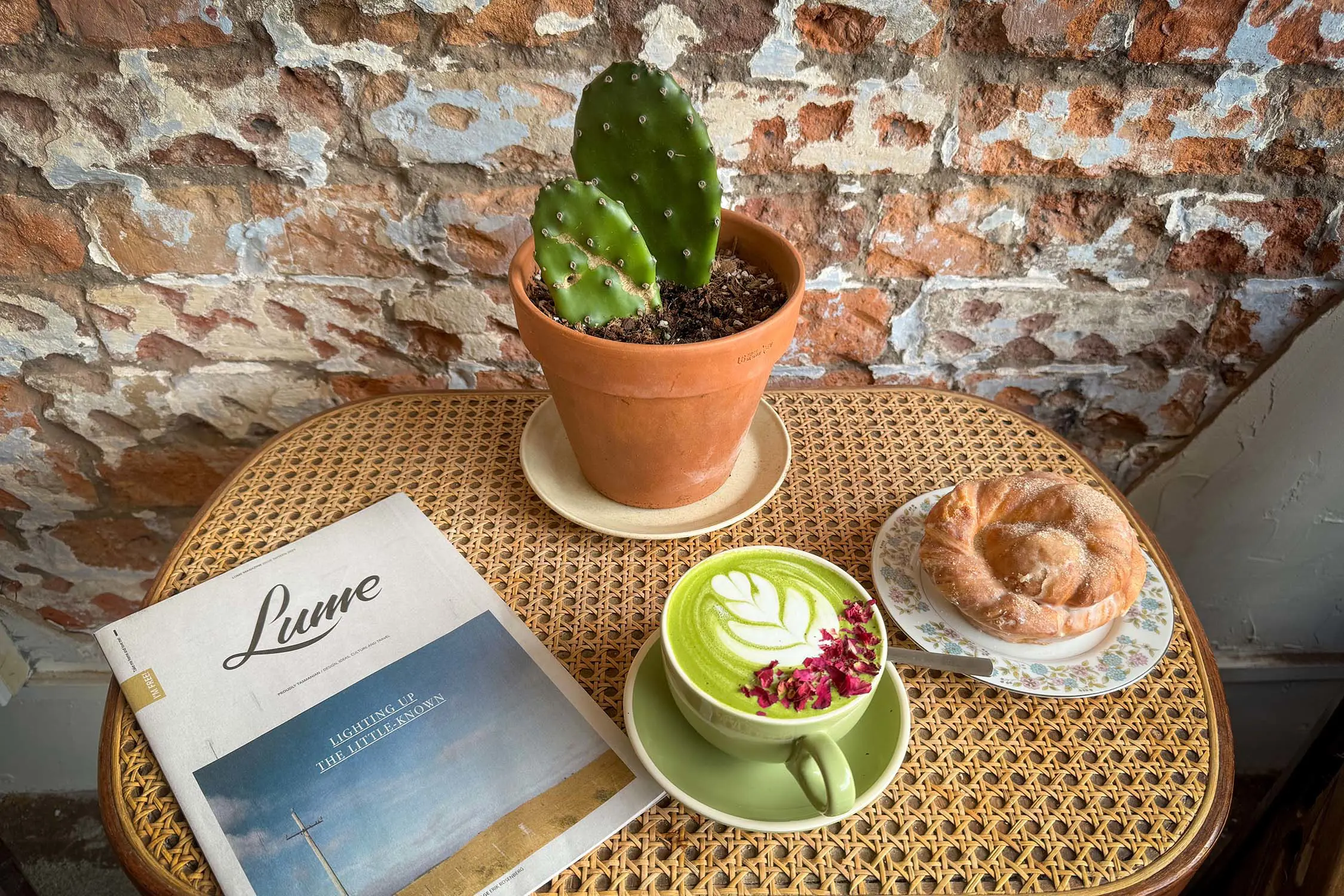 A small wicker table against a rustic brick wall, holding a cactus pot plant, a pastry, a matcha latte and a copy of Lume magazine.