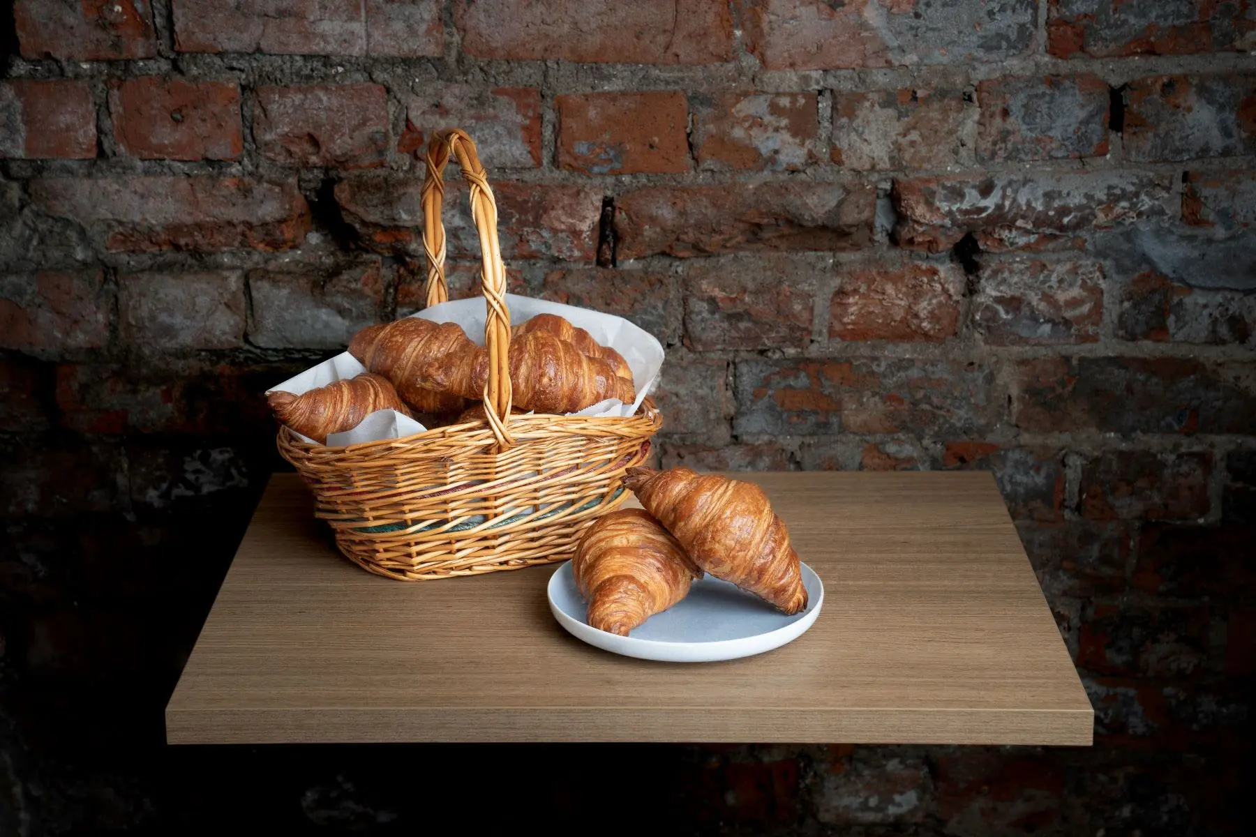 A basket of croissants and two more on a plate sit on a small wooden side table in front of a rustic brick wall.