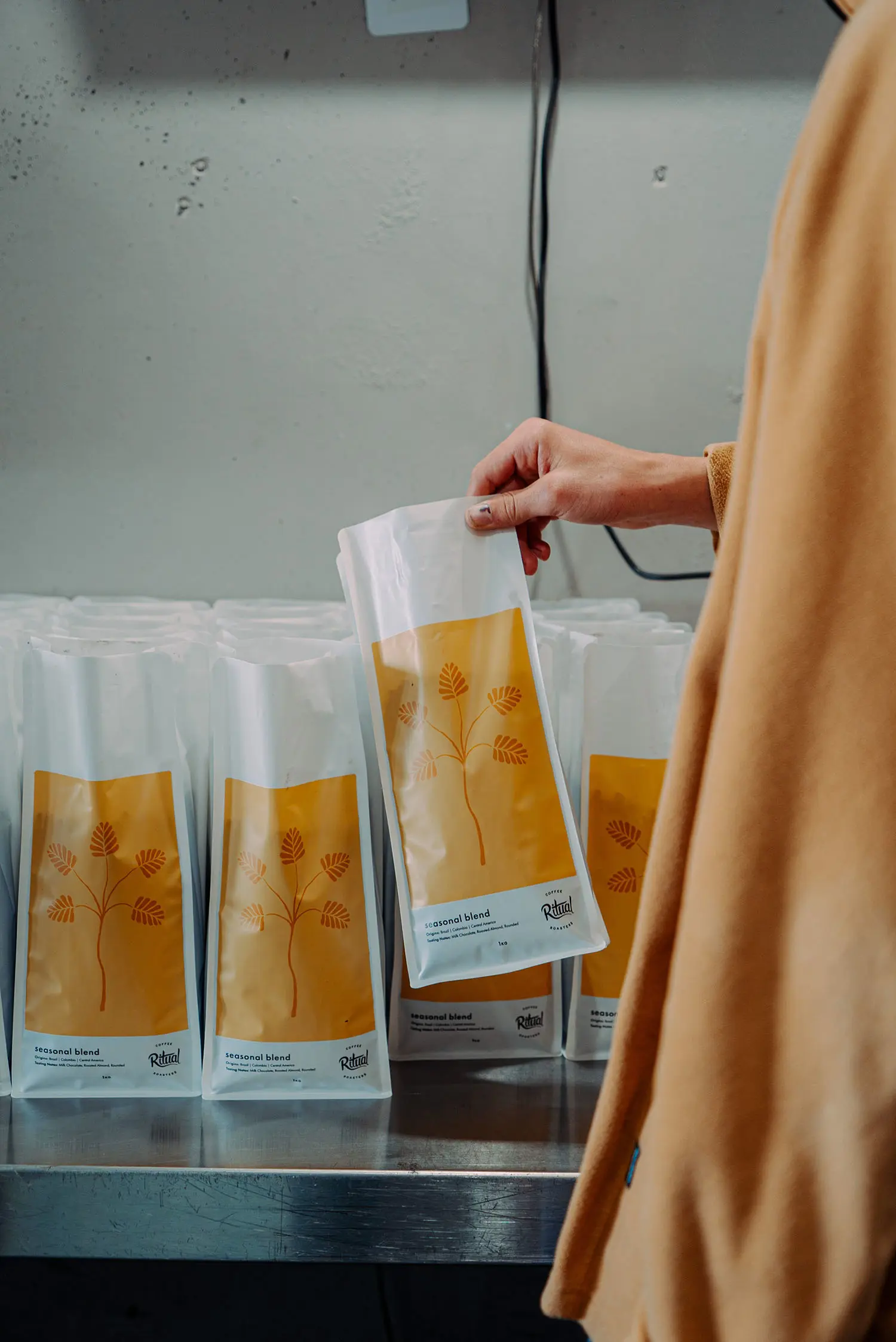 A person picks up a bag of coffee from rows of bags on a silver benchtop. The white bag has a yellow and orange leaf image and the label reads 'Seasonal Blend'.