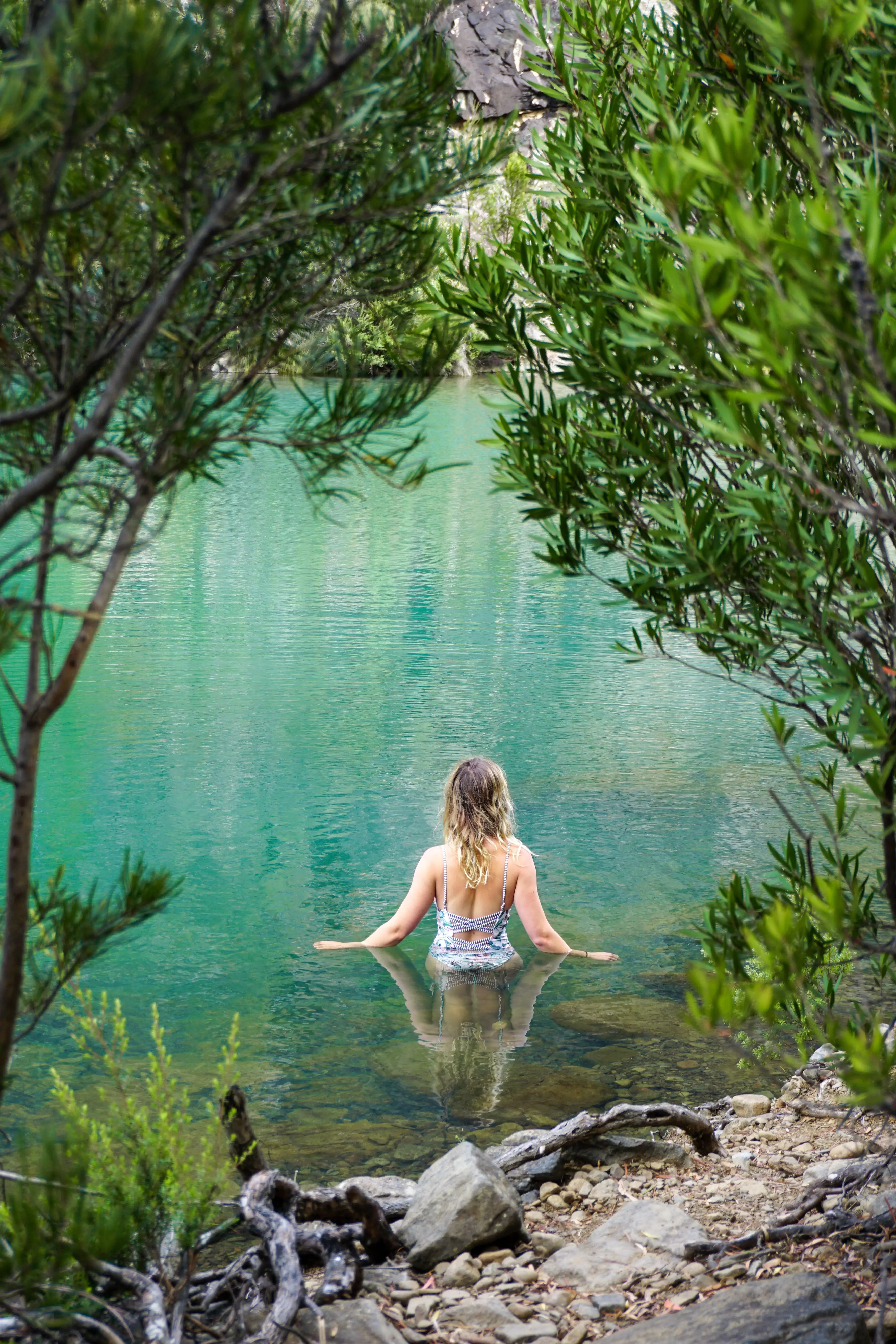 Framed by green leafy trees, a woman in a swimsuit is waist-deep in a natural turquoise waterhole, gazing with her back to the camera towards the water.