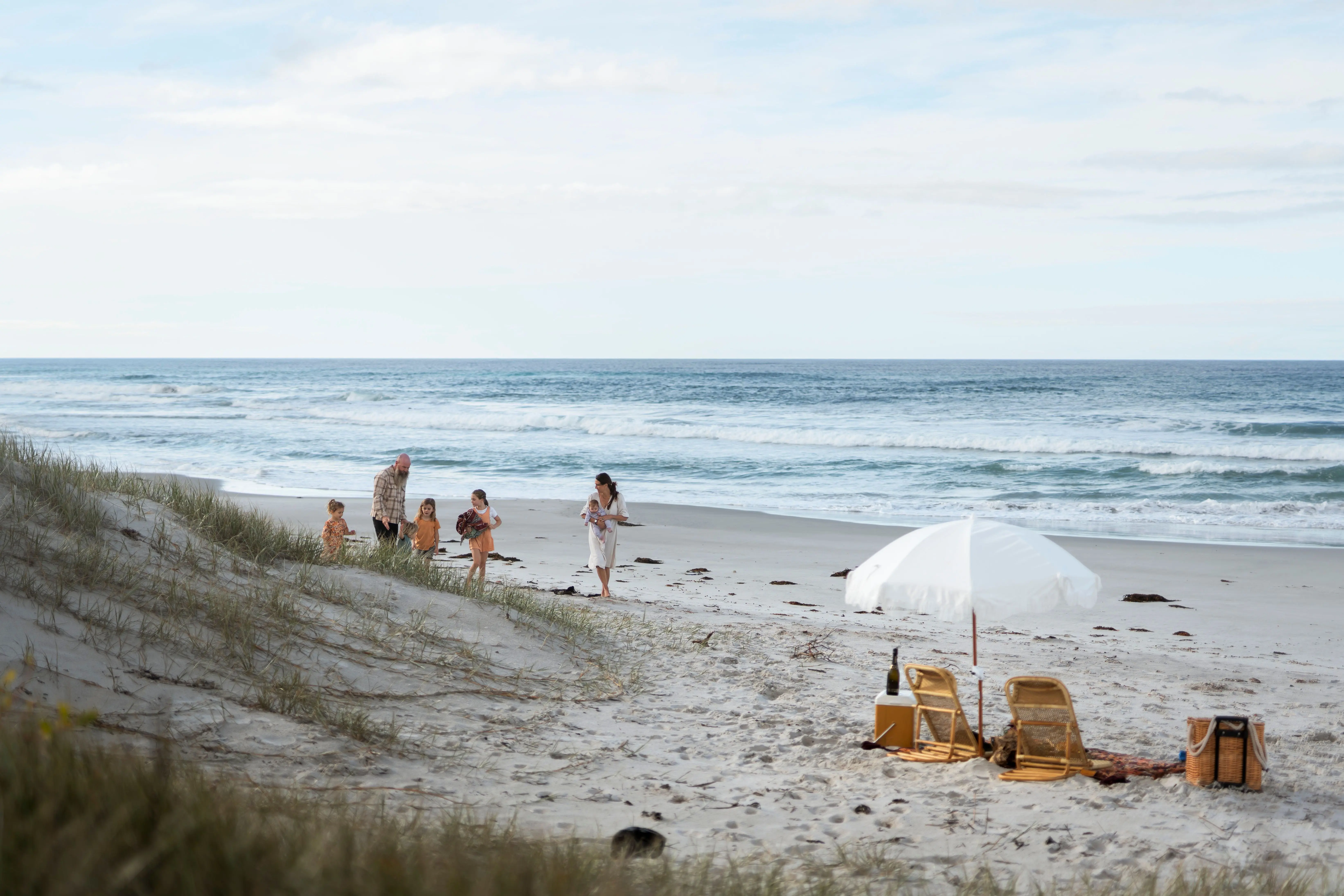 A mother carrying a baby and a father with three daughters on a white-sand beach walk towards a white umbrella picnic setting.