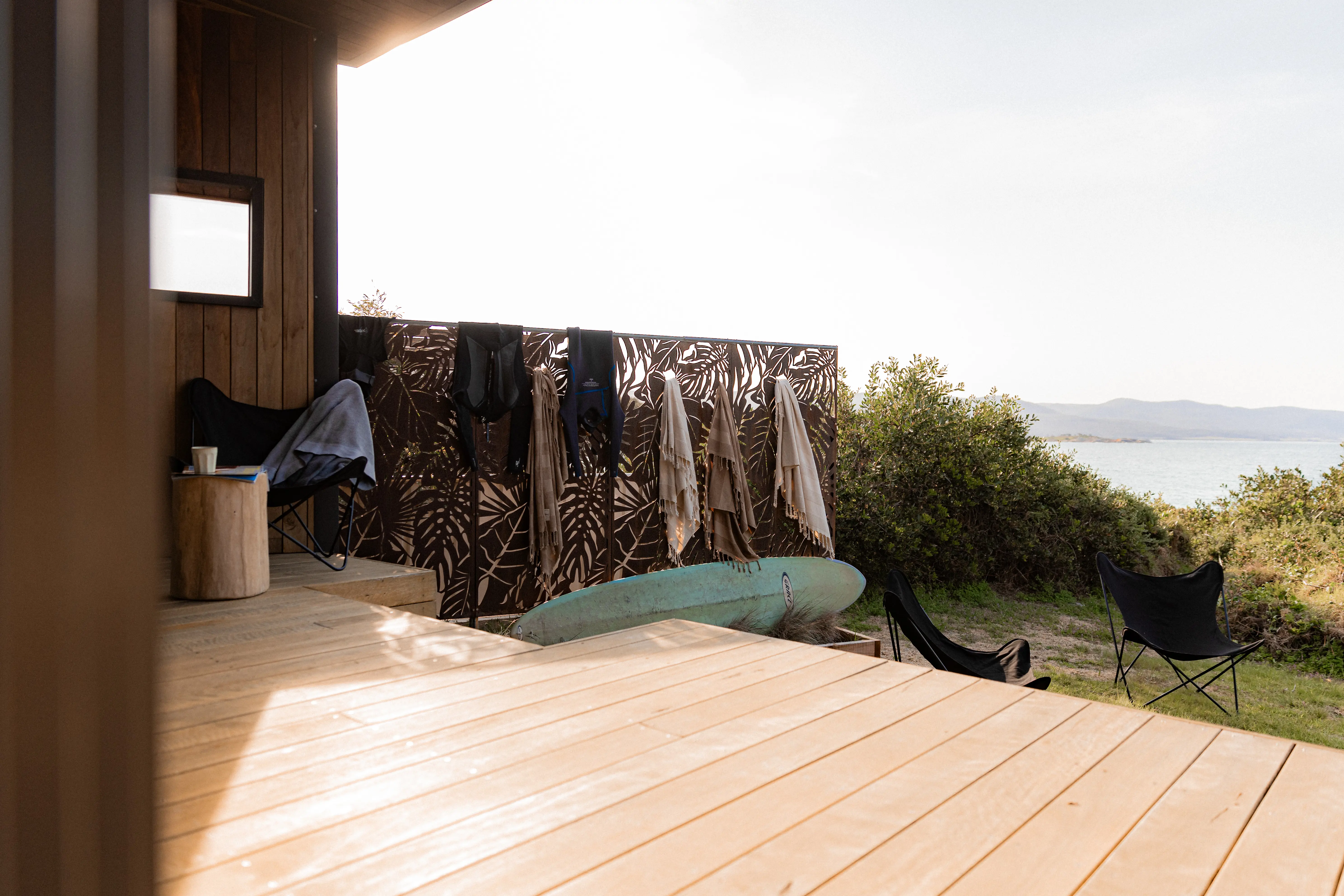 Wooden coastal deck beside a cabin, with beige towels and black wetsuits hanging on a decorative brown privacy screen, an aqua-coloured surfboard resting below, and two black outdoor chairs overlooking the ocean and distant hills.