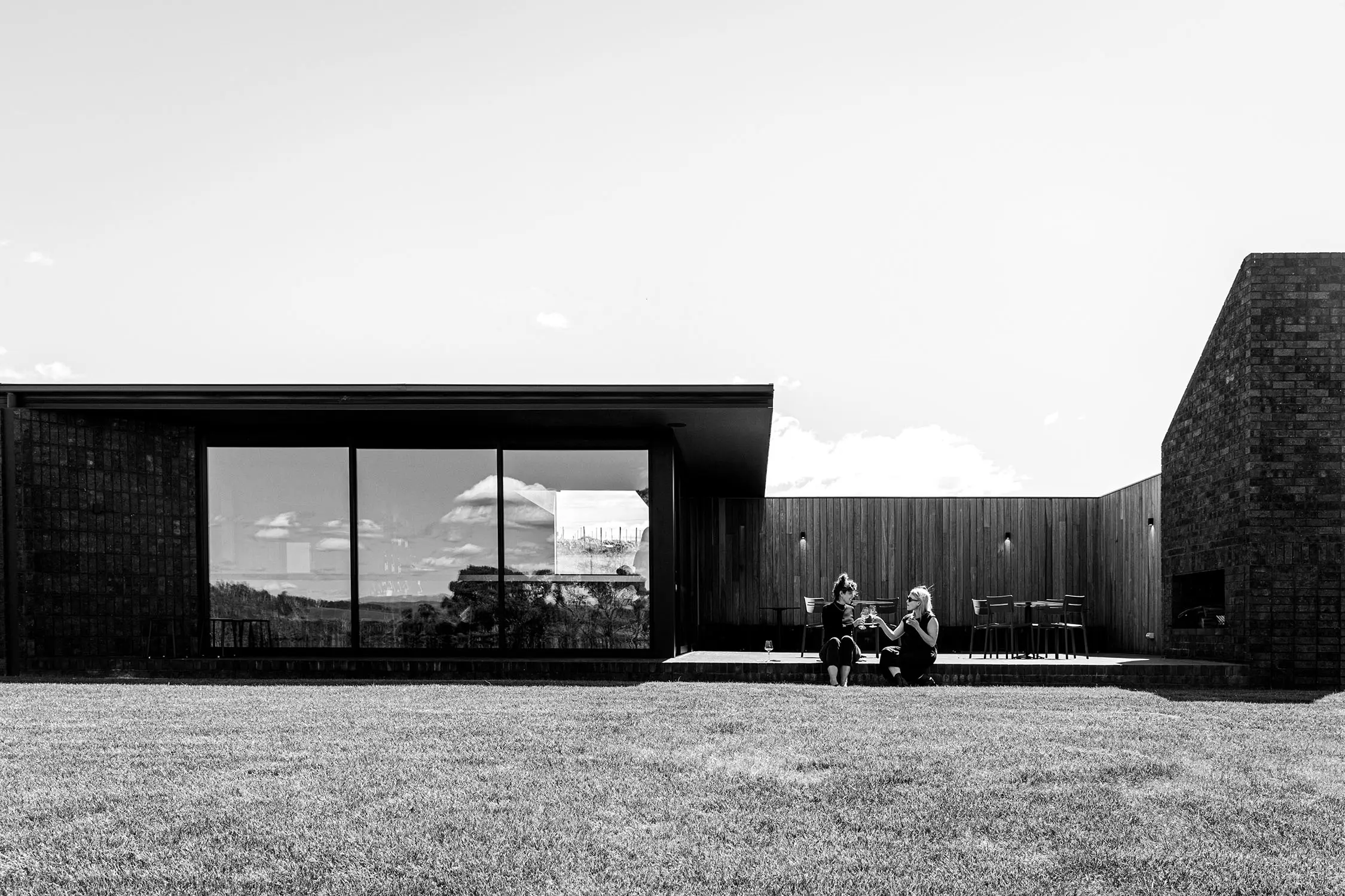 The outdoor facade of a stunning architectural and angular winery, with dark wood cladding and full-length windows. Two people sit on the deck drinking wine.