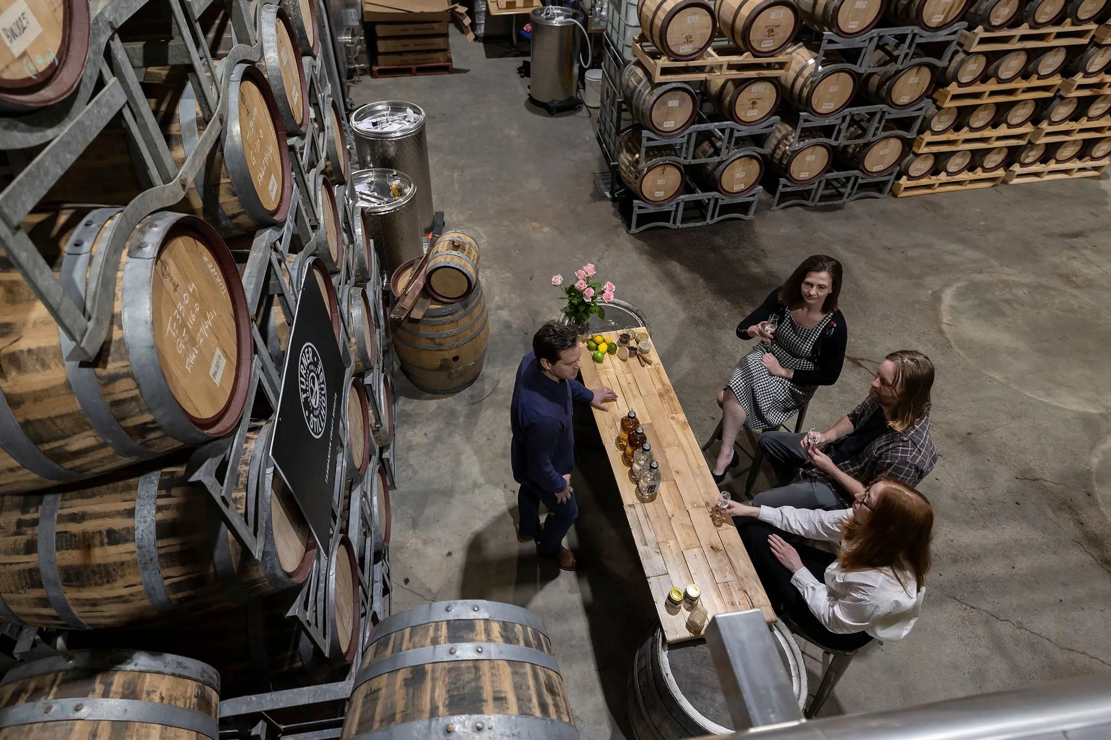 A top-down view of a large warehouse-style whisky distillery. Three people sit at a bar doing tastings, surrounded by huge stacks of barrels.