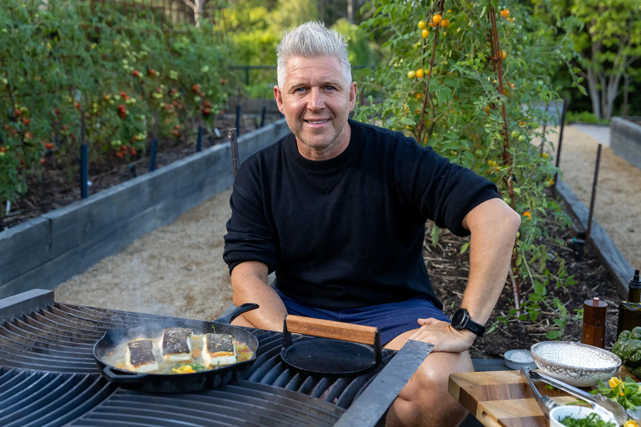 A man in a black shirt sits among raised garden beds, at a barbecue. On the grill is a pan with fillets of fish frying.