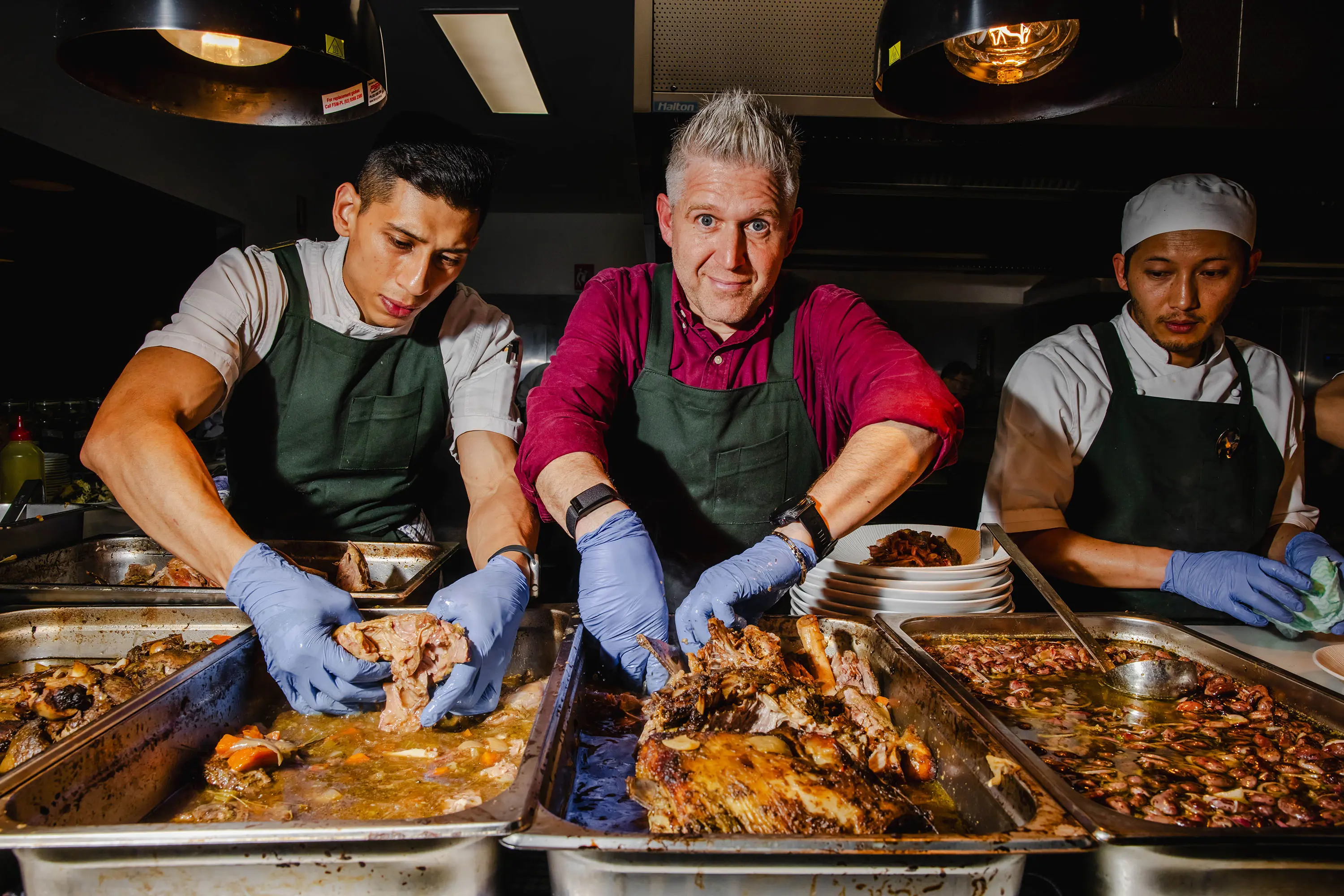 Three chefs wearing aprons stand over a row of huge bain marie trays, filled with delicious fall-apart meat and pan juices.
