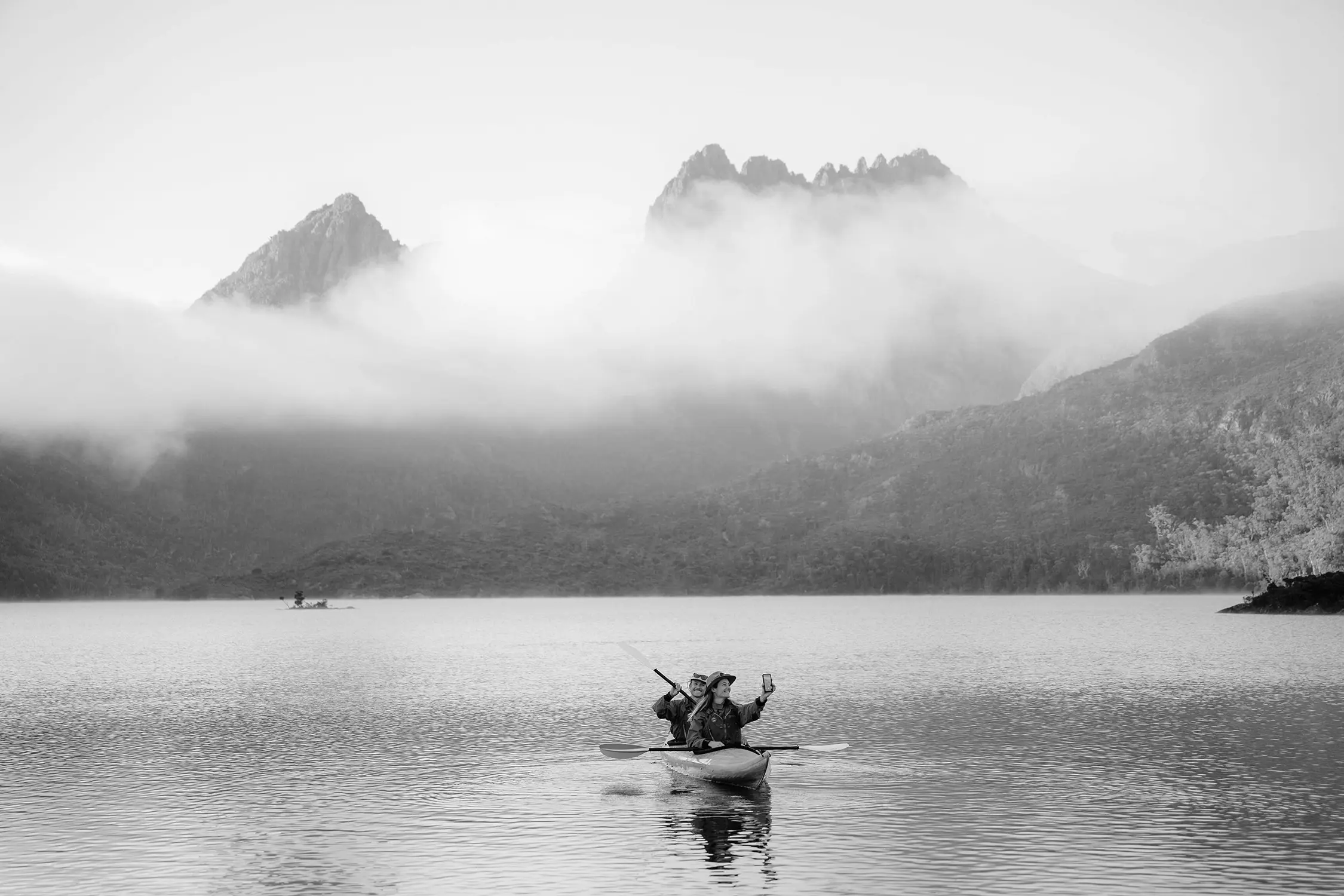 Two people paddle in a small kayak on a dappled lake surrounded by mountains. Out of the fog rises sharp peaks behind them.
