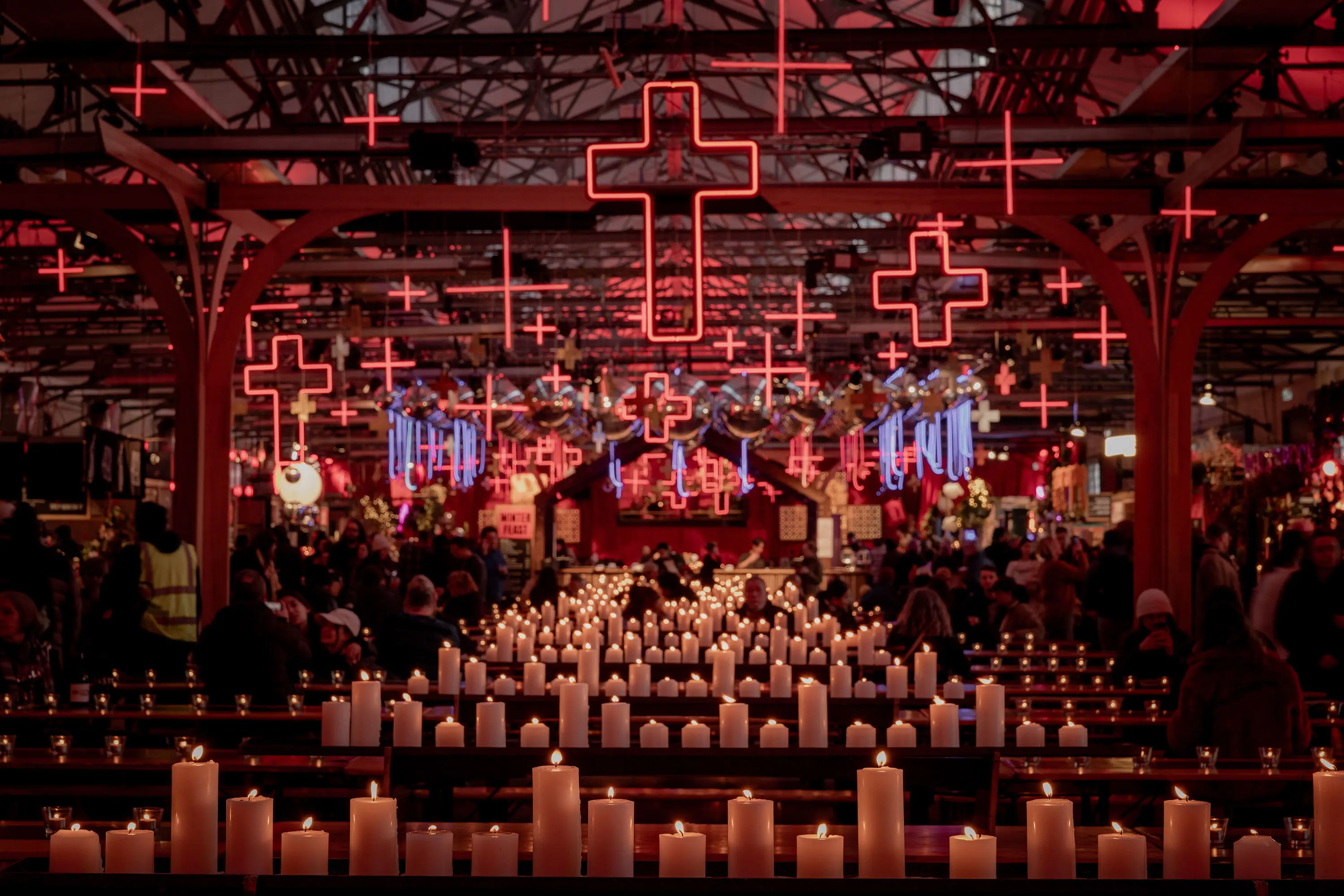 A crowded venue filled with people, illuminated by glowing red crosses hanging from the ceiling and lined with candles on the tables.