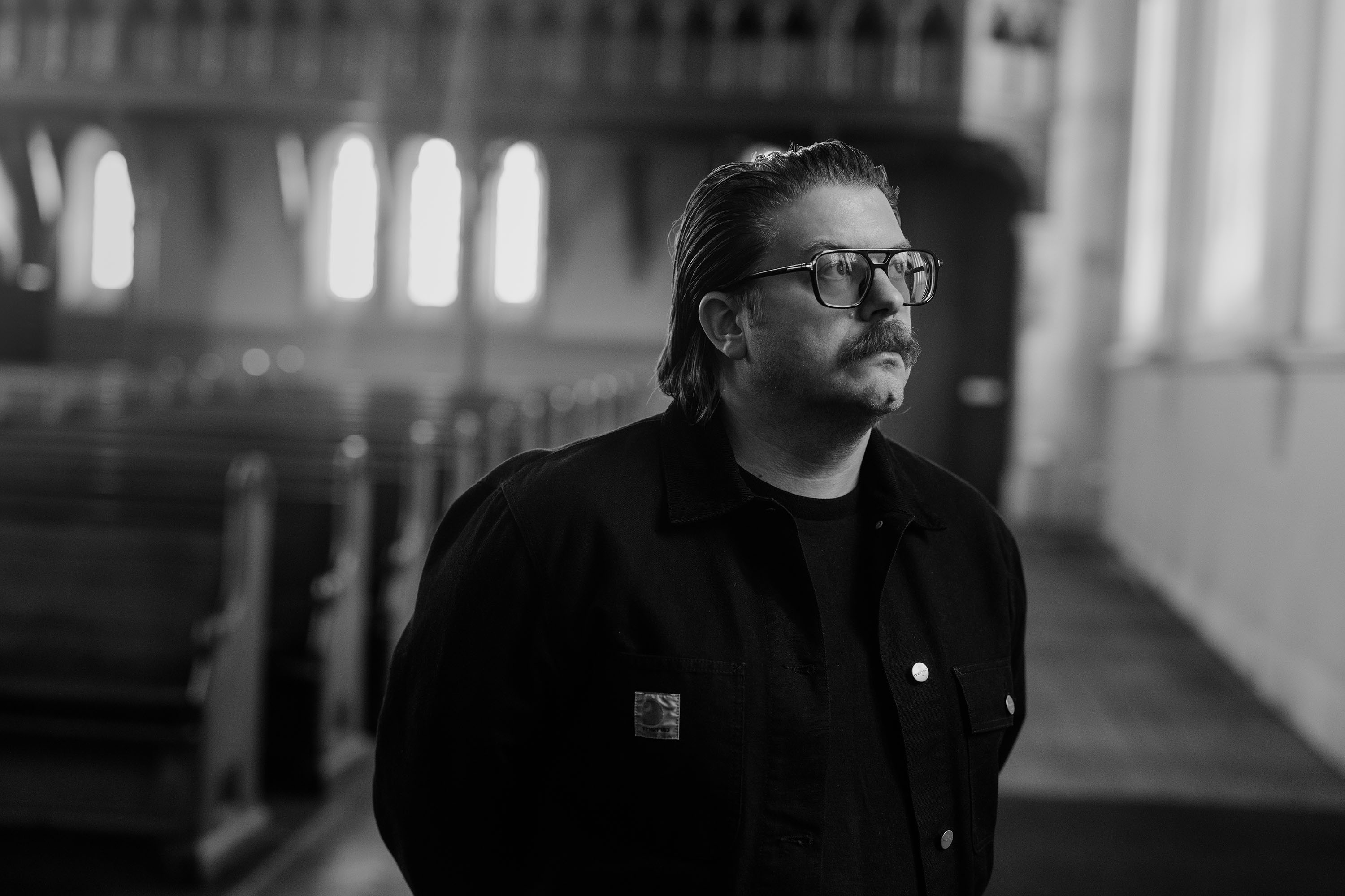 A man wearing a black jacket, large glasses with a moustache and slicked-back hair stands gazing out a window. Behind him, the out-of-focus background of a church with pews and sculptural windows is visible.
