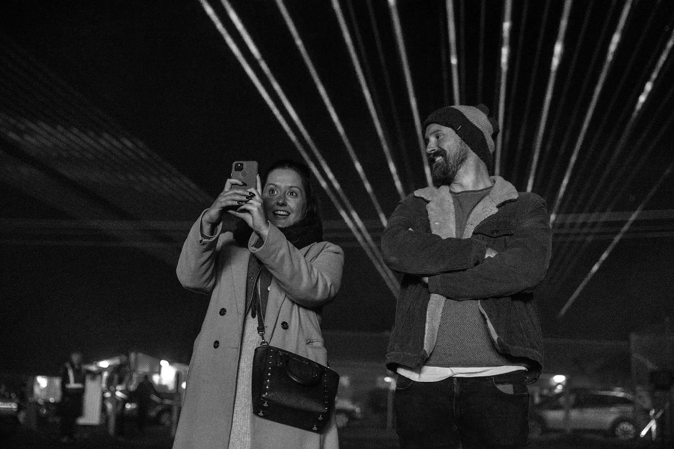Two people, dressed for cold weather, stand outside with a laser light-show radiating beams behind them. The woman holds up a phone to take a photo.