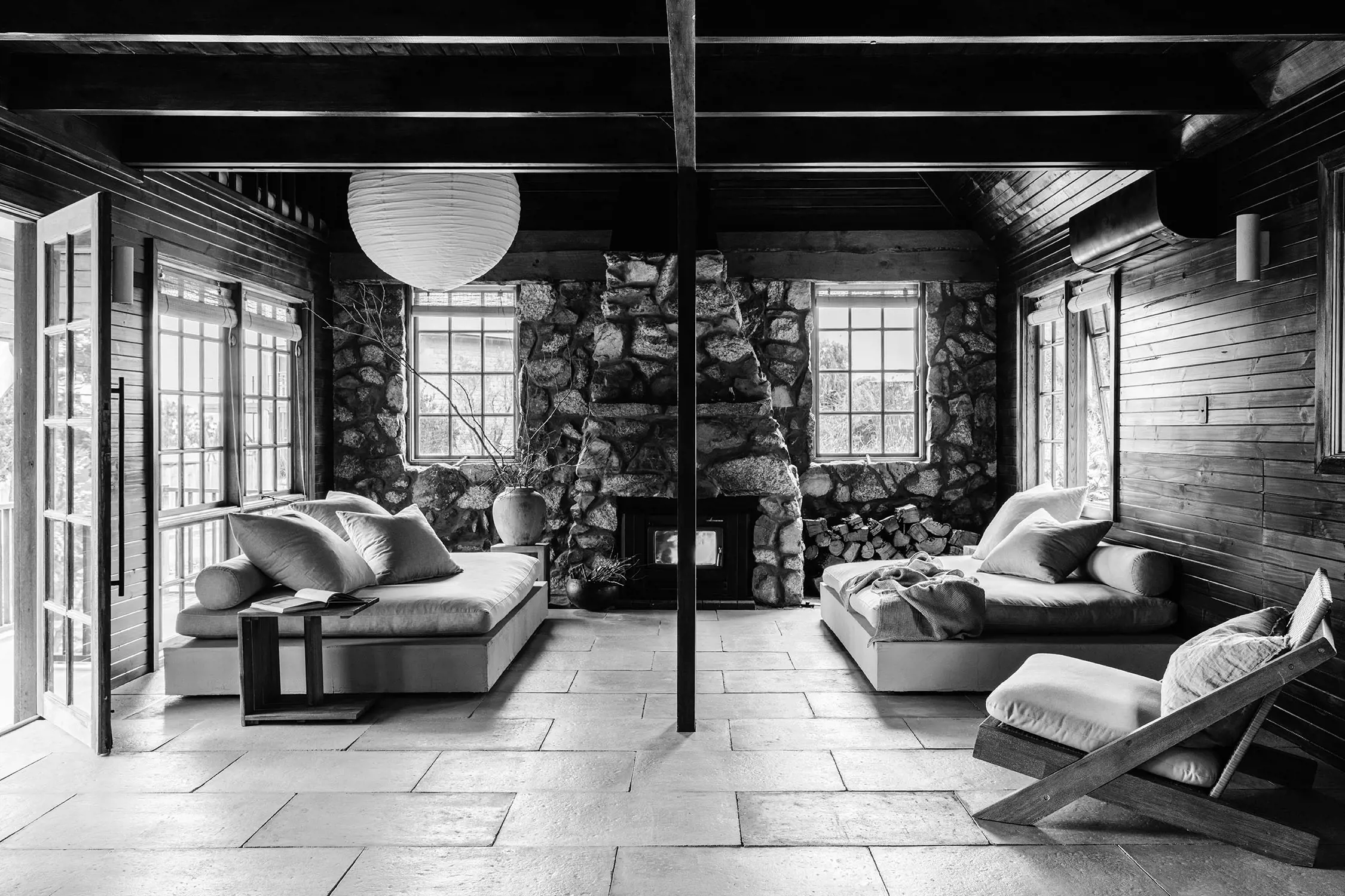 A black and white image of a rustic living room with two large windows, a stone fireplace, and two cushioned lounge chairs. The room has wooden walls and beams, a tiled floor, and a round paper lantern hanging from the ceiling.