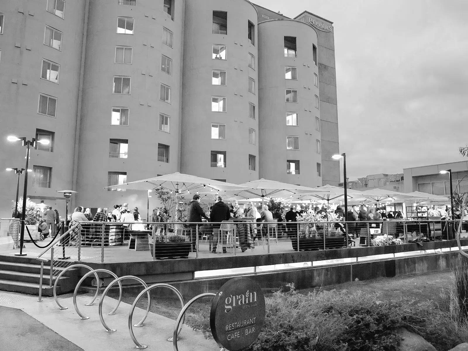 A black and white evening scene captures diners gathered under white umbrellas at an outdoor restaurant beside towering heritage grain silos. The contemporary terrace showcases industrial architecture transformed into a vibrant winter dining experience.