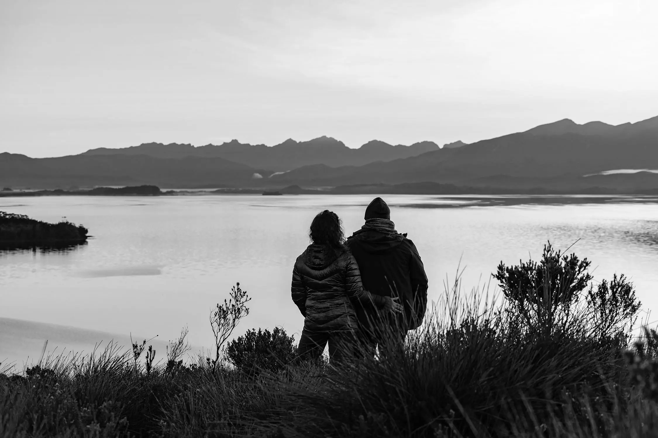 Two people in winter jackets stand among native grasses and shrubs, facing calm water with a mountain range visible across the bay.