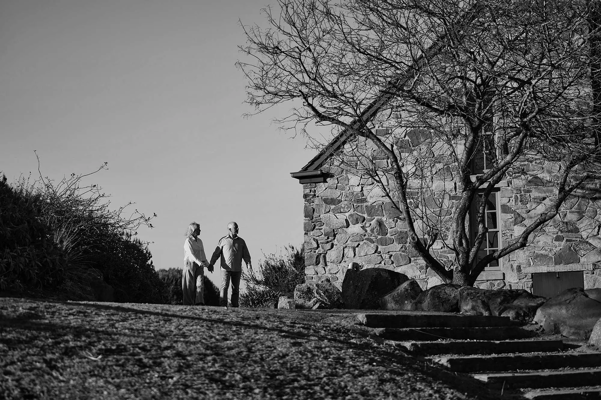 Two people hold hands while walking on a gravel pathway near a heritage stone building. Bare winter trees and stone steps frame the scene.
