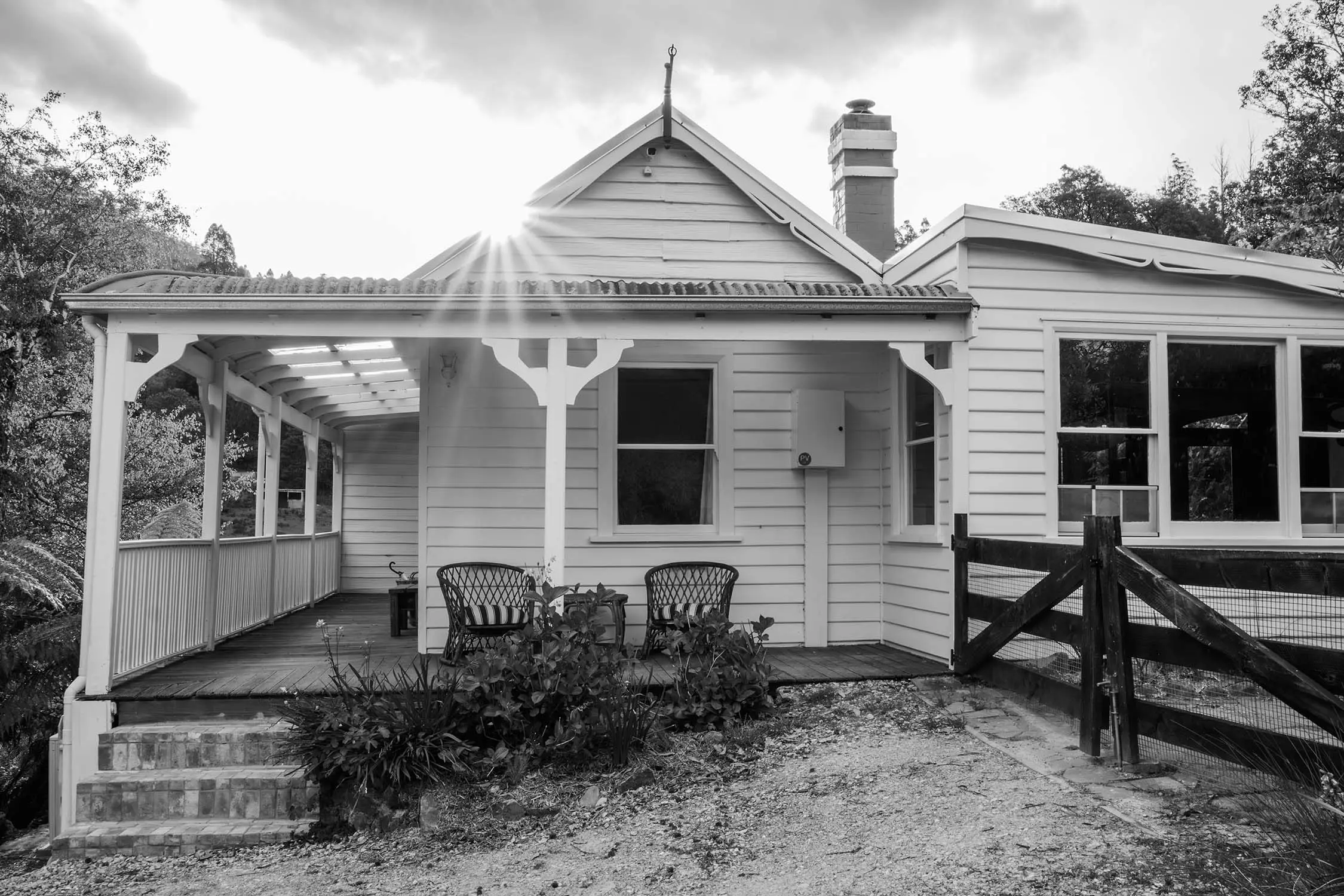 White weatherboard cottage features decorative trim and a brick chimney. Two wicker chairs sit on the covered verandah with sun flare above the roofline.
