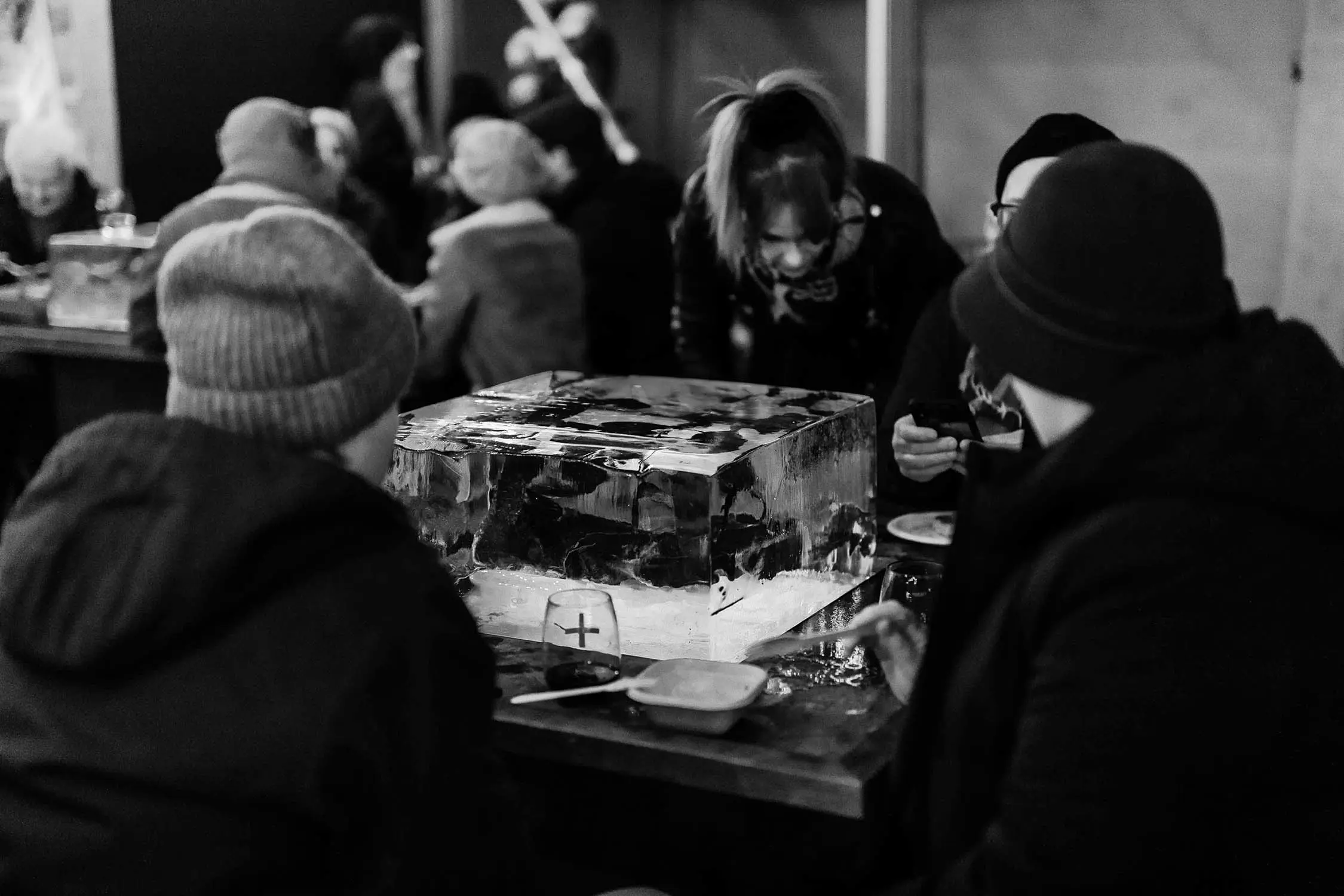 A black and white photograph shows people seated around a large rectangular ice block on a table. The ice sculpture serves as a centerpiece with glasses and plates arranged around it in dim lighting.