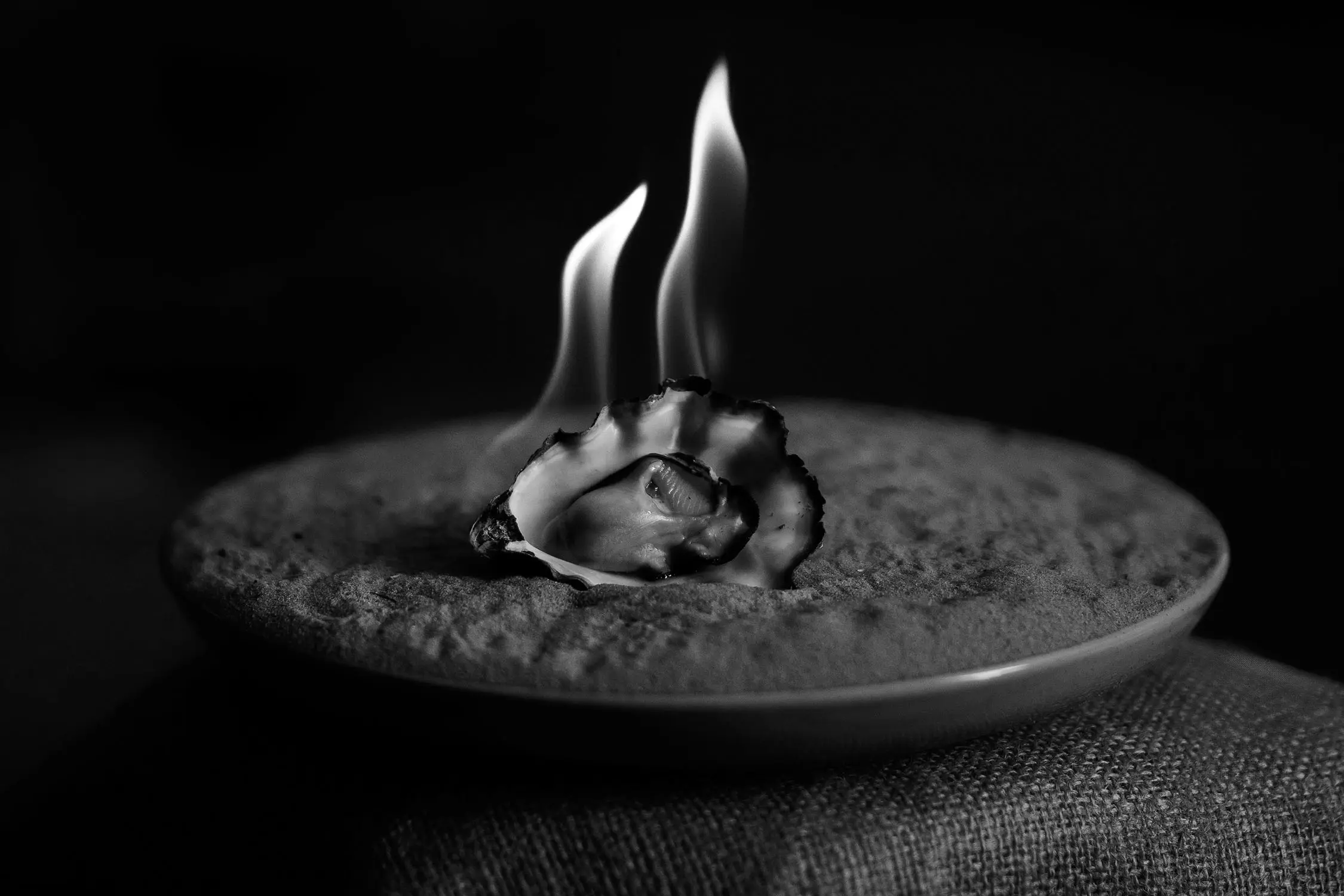 A black and white close-up shows a fresh oyster with flames rising from it, placed on a textured ceramic plate. The fire creates dramatic lighting against the dark background.