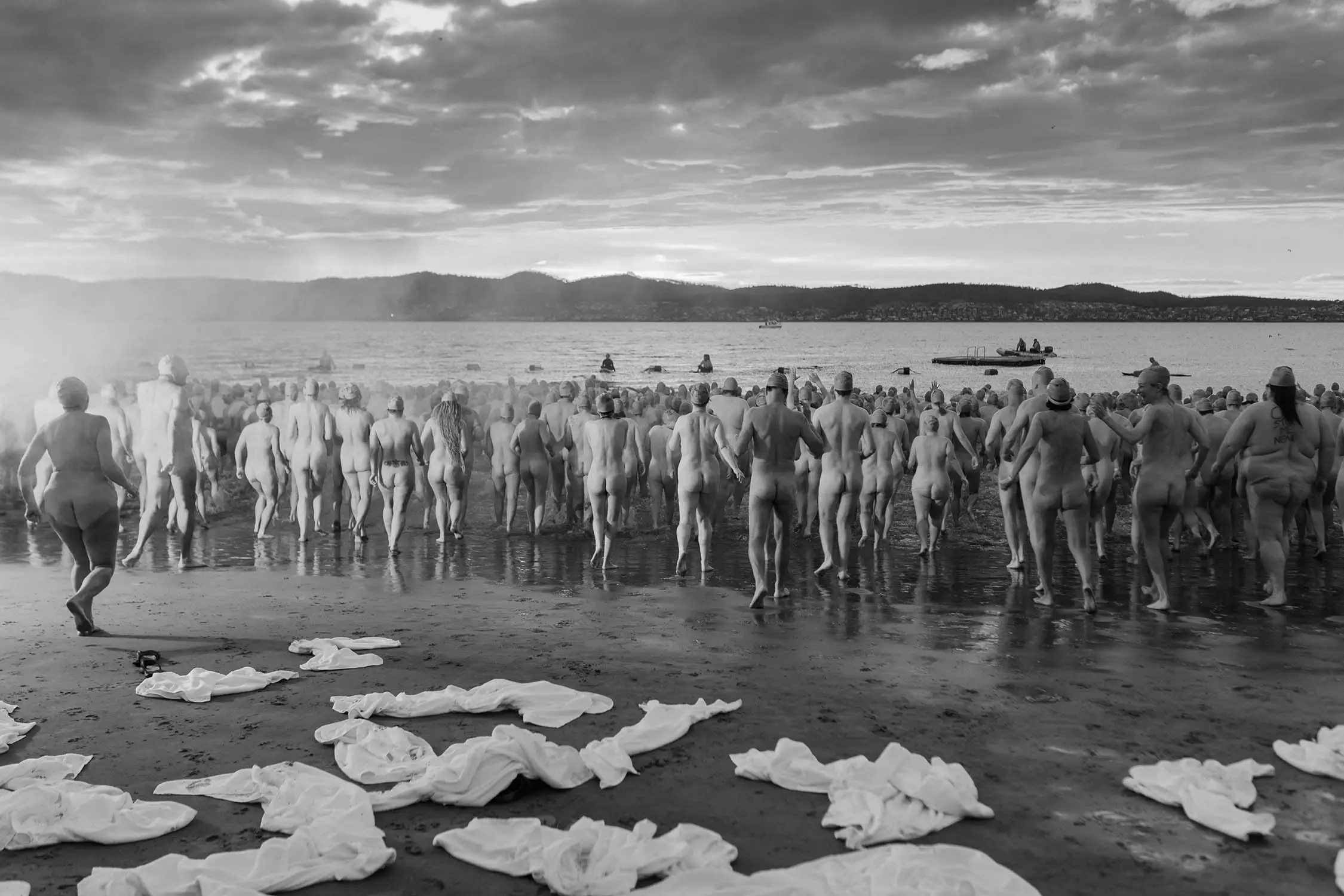 A huge crowd of people, naked except for swimming caps, run into the water. White towels are strewn on the sand behind them.