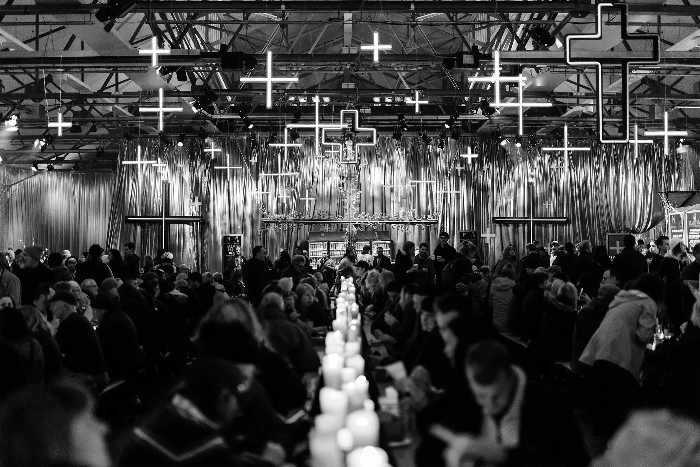 A crowded venue filled with people, illuminated by glowing crosses hanging from the ceiling and lined with candles on the tables.