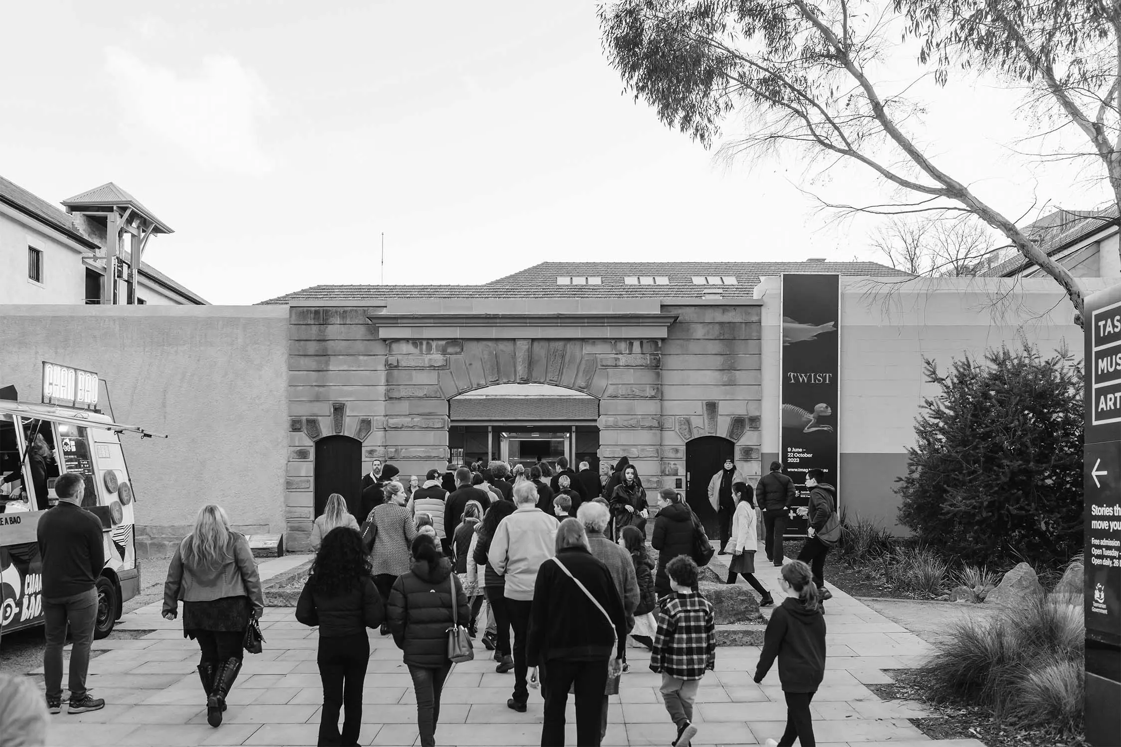 A black and white photograph depicting a crowd of people walking towards an entrance framed by a large stone archway. The scene includes a food truck on the left side, and various buildings can be seen in the background. Some individuals are wearing winter jackets as they approach the entrance, where others are already gathered. Trees and shrubs are visible on the right side, along with a nearby sign. The atmosphere suggests a busy event or gathering.