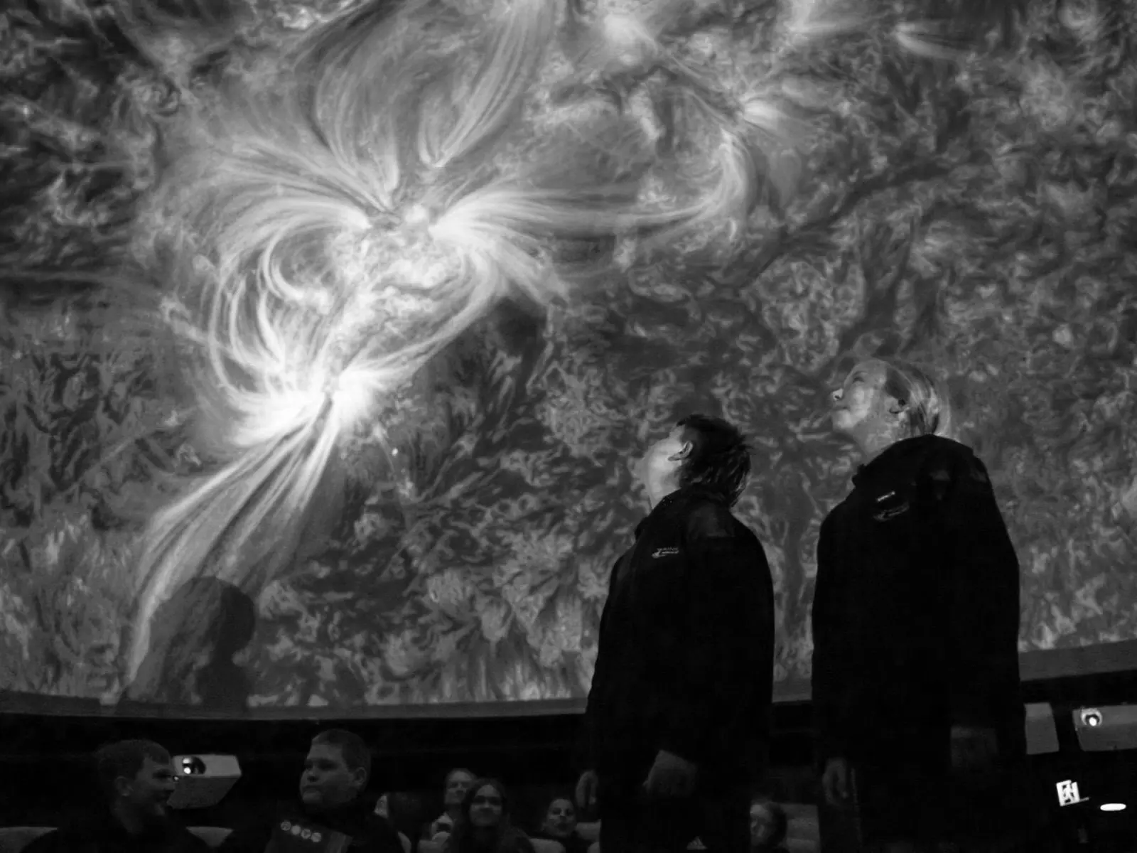Two people stand inside a domed planetarium, the swirling space projections covering their faces and the ceiling above.