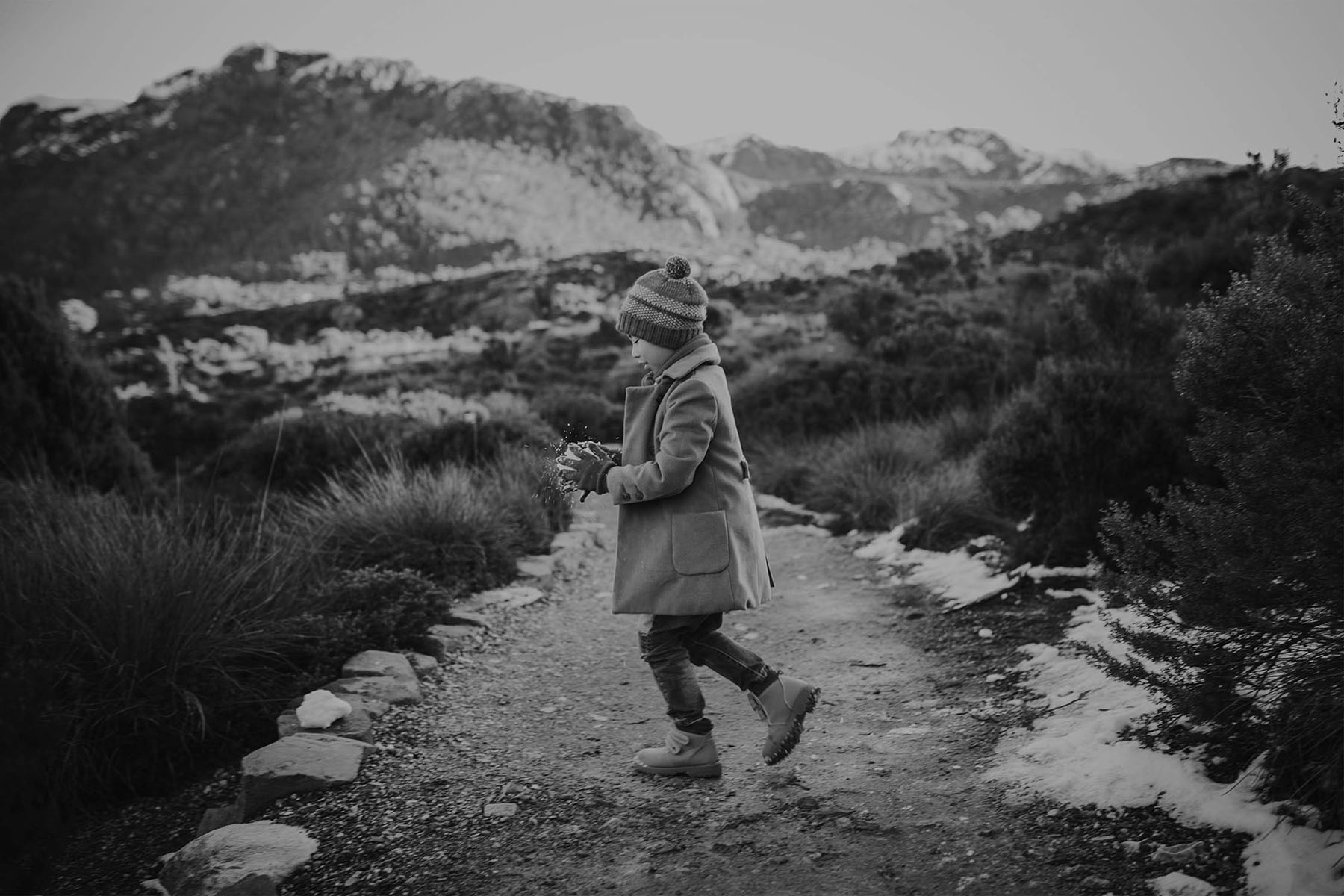 A child in winter attire walks along a path, tossing snowflakes in the air with mountains in the background.