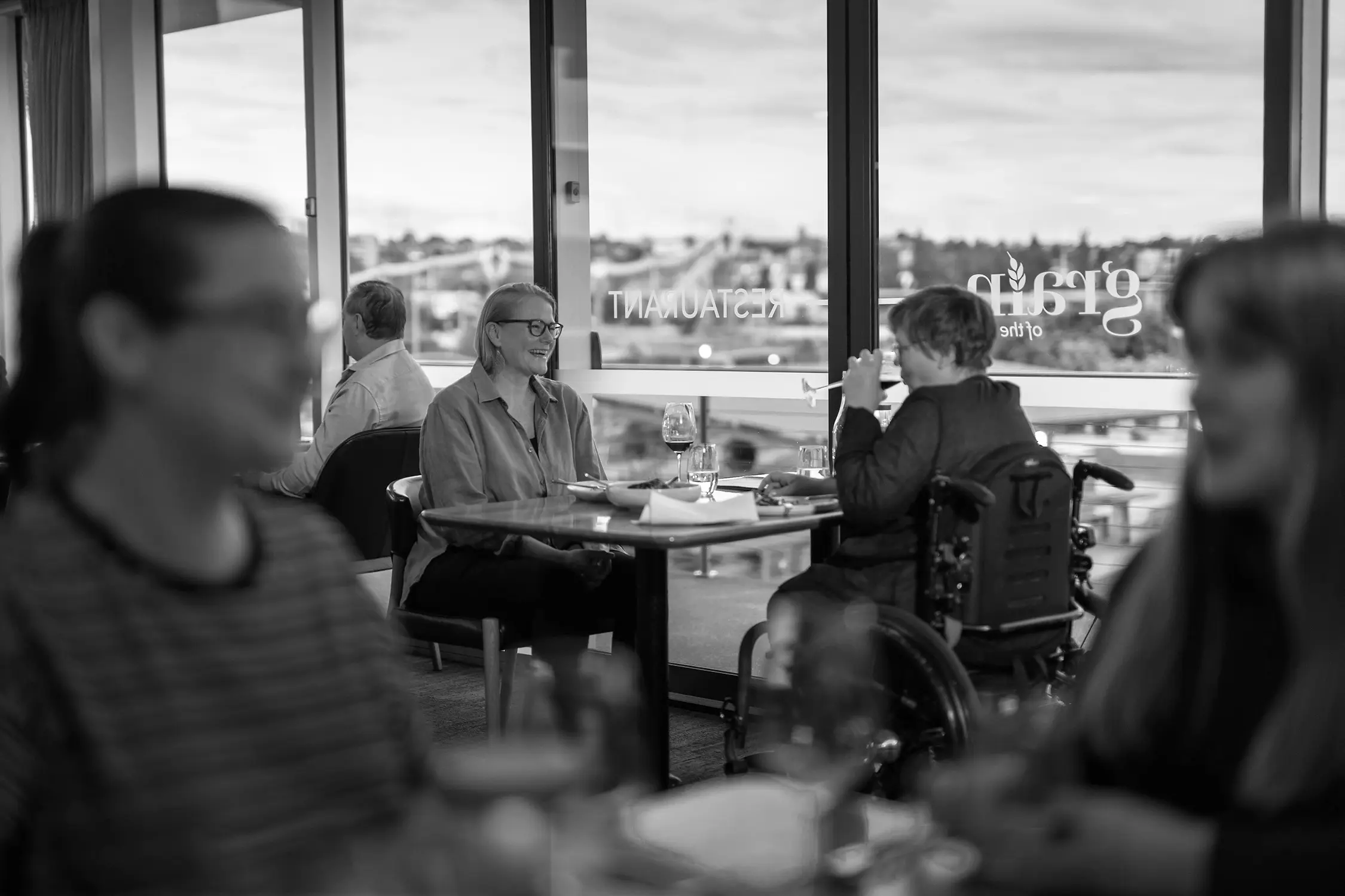 Two people, one in a wheelchair, sit at a table against large glass windows at the edge of a busy restaurant.