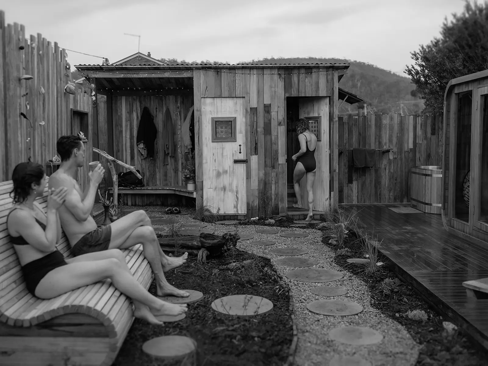 Two people sit on a sculptural wooden bench in a rustic outdoor courtyard. A third person is entering the wooden sauna at one end, the pathway to it lined with stepping stones.
