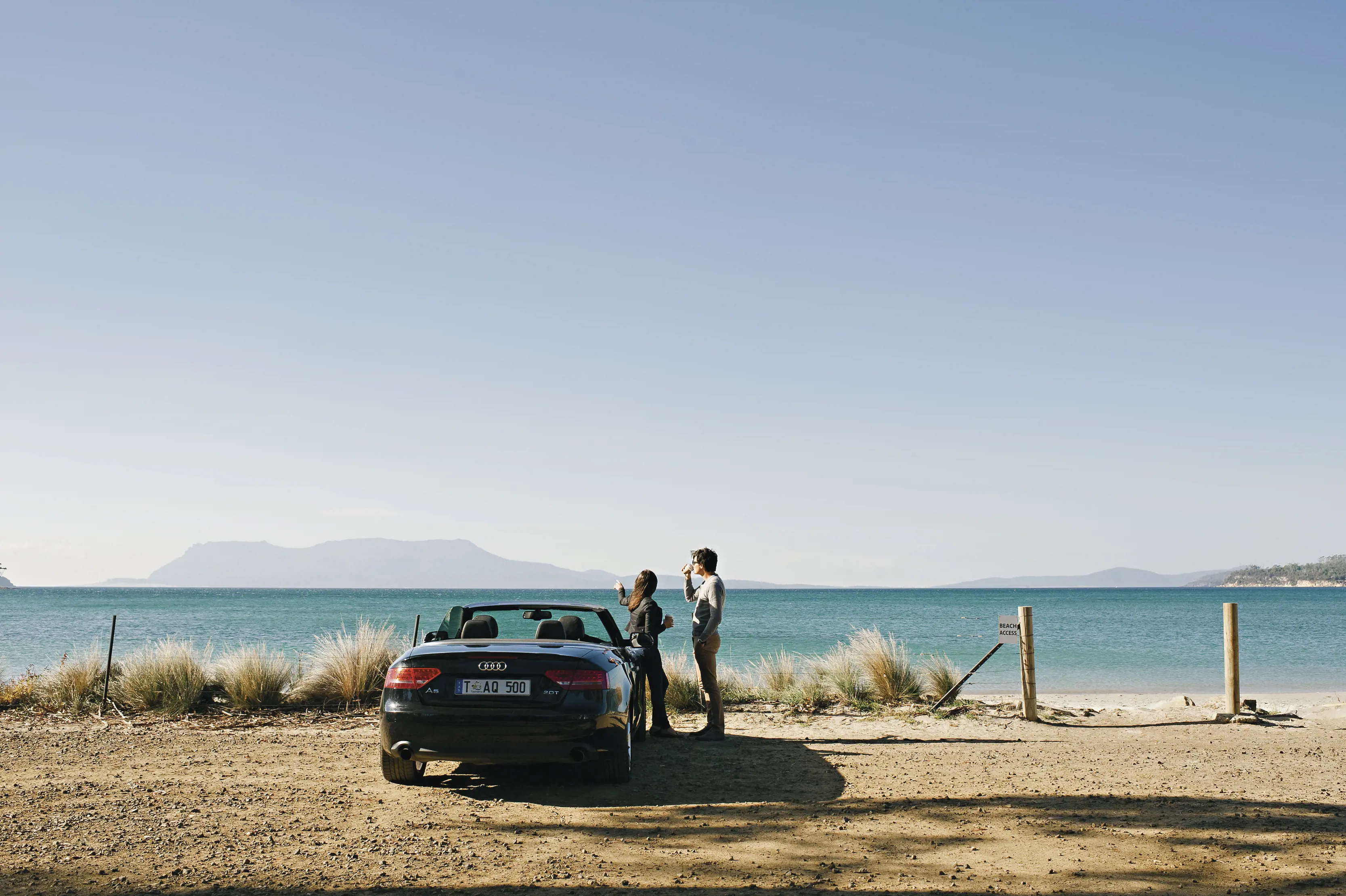 Two people standing next to a convertible car at Spring Beach, Orford. You can see Maria Island in distance.
