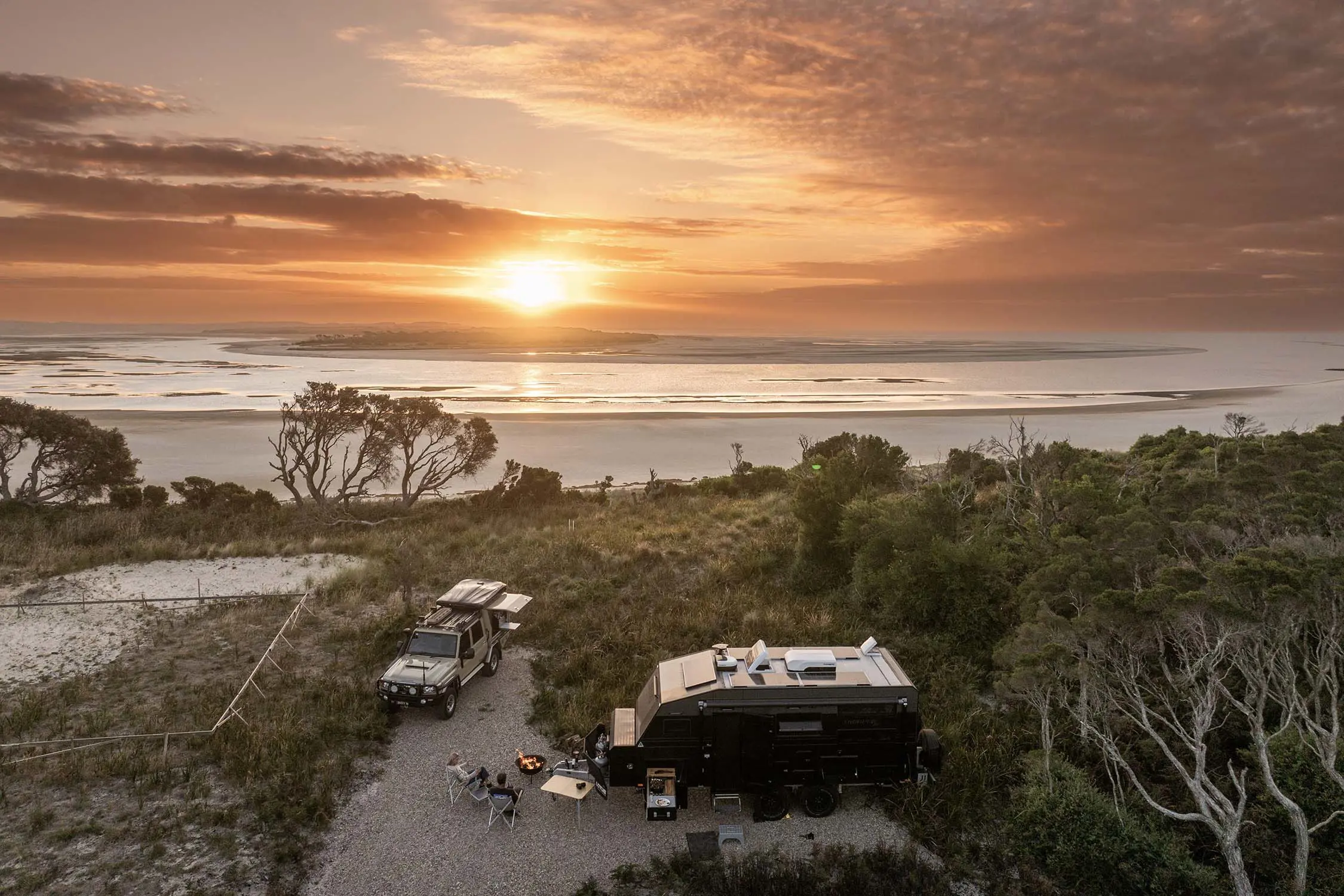 An aerial view of a beachside campground, where a caravan and 4WD are parked, people relaxing outside them. The sun sets over the beach in the background.
