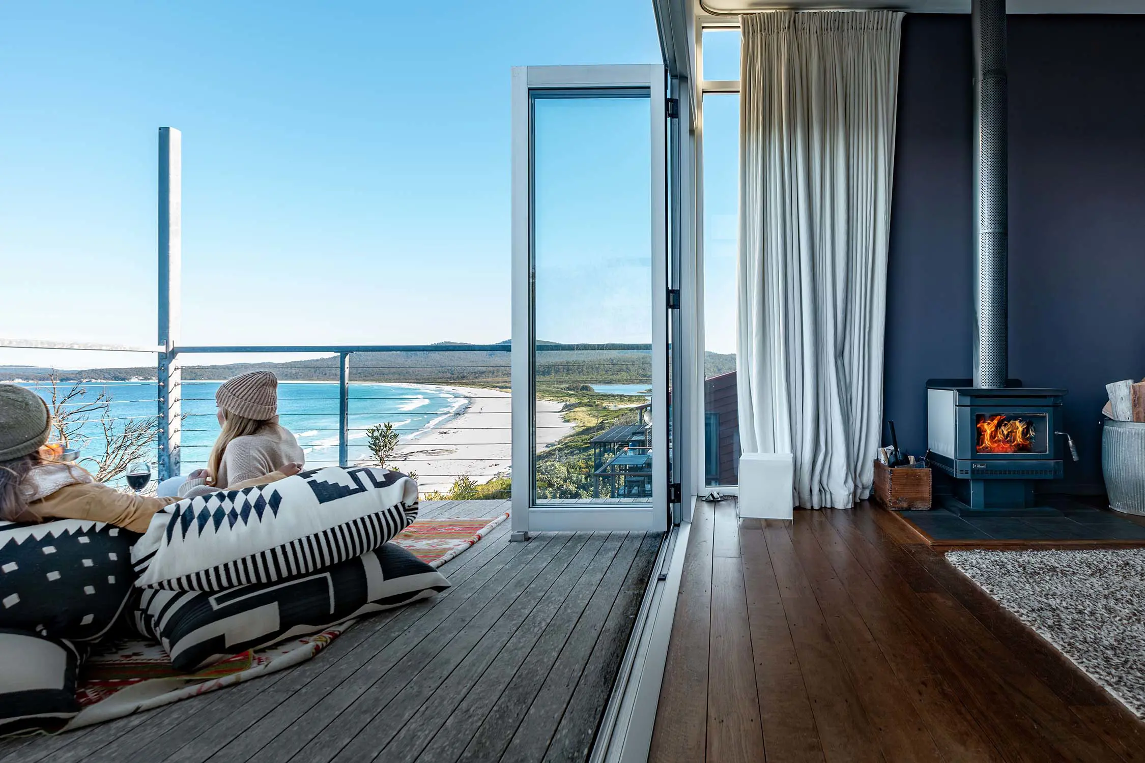Two people sit in beanbags on an outdoor deck, with the wall-to-ceiling glass doors pulled wide open, revealing the fireplace inside.