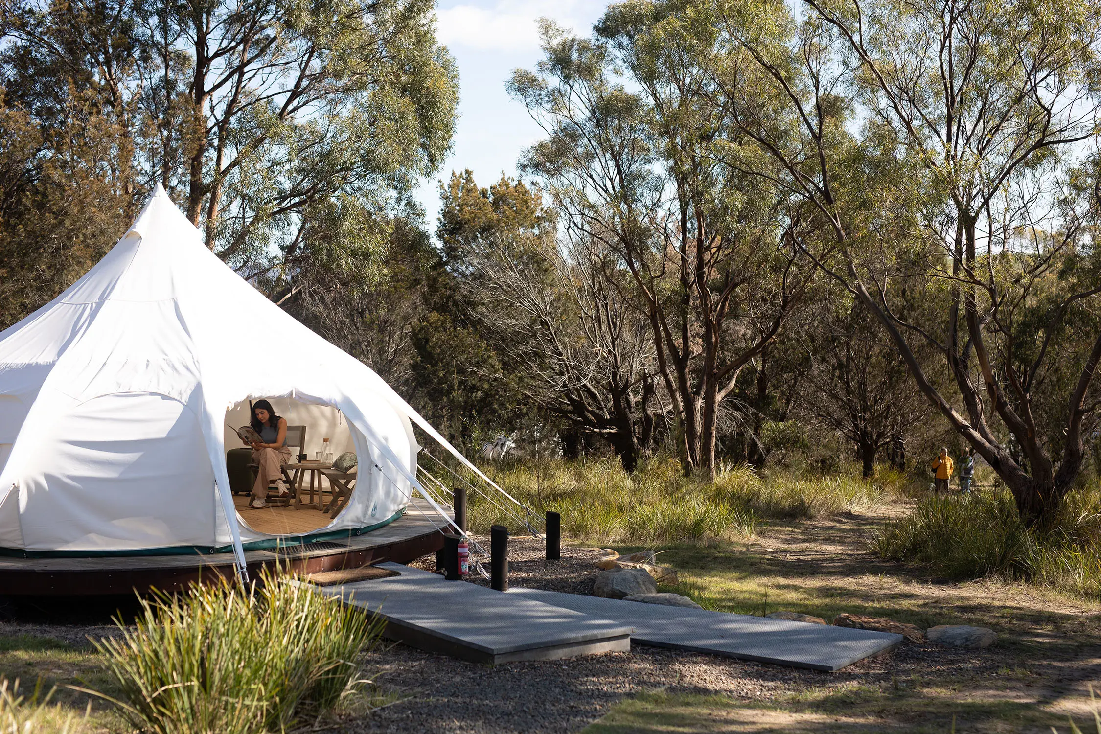 A woman sits inside a white glamping tent, nestled on a wooden platform amongst the bush.