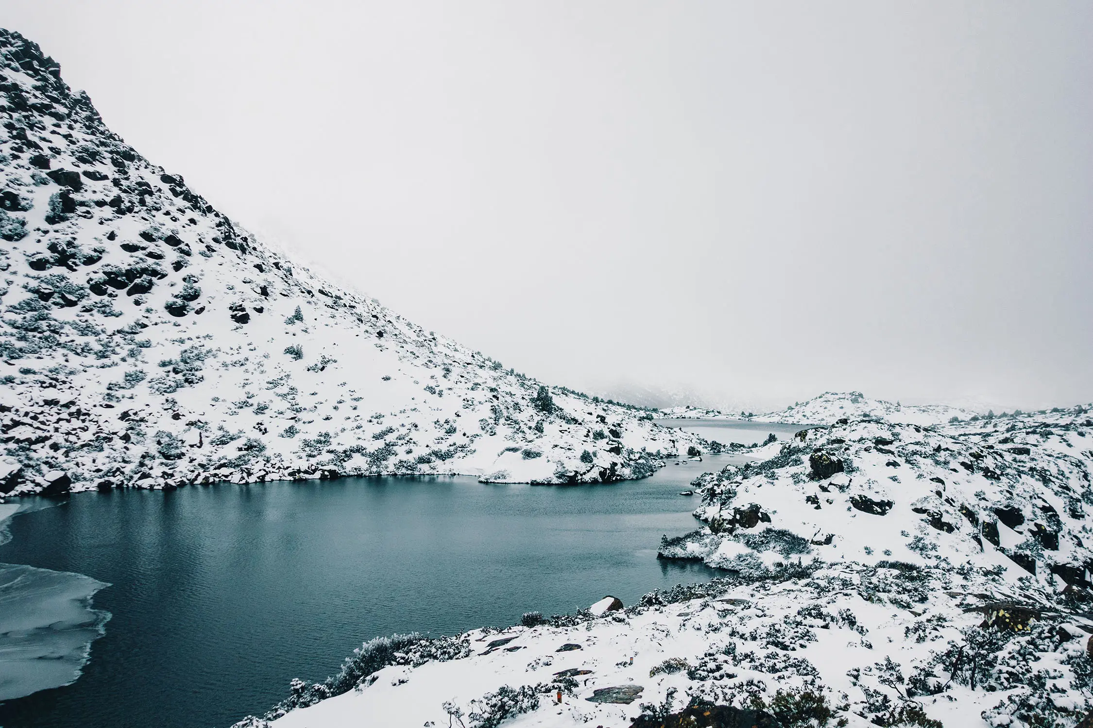 A stark, snow-covered landscape surrounding a dark alpine lake. The sky is completely filled with heavy white cloud.