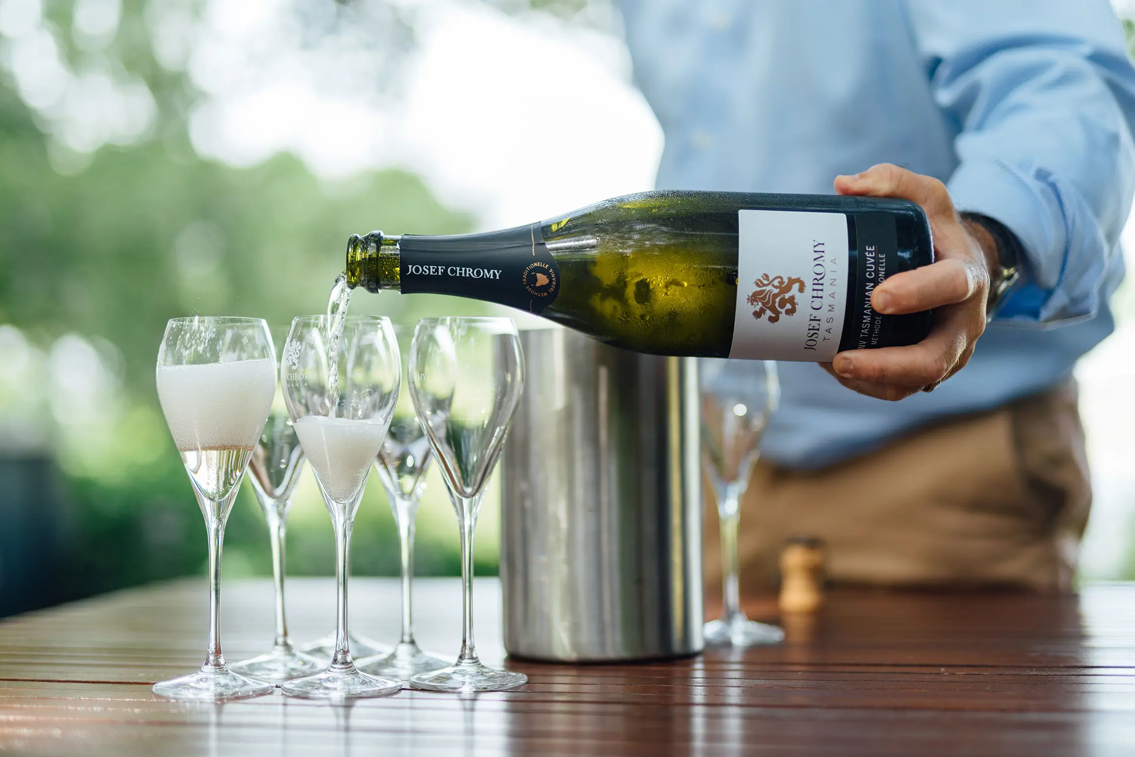 At an outdoor wooden table, a person artfully pours a bottle of sparkling wine into five champagne flutes. The label on the bottle reads 'Josef Chromy'.