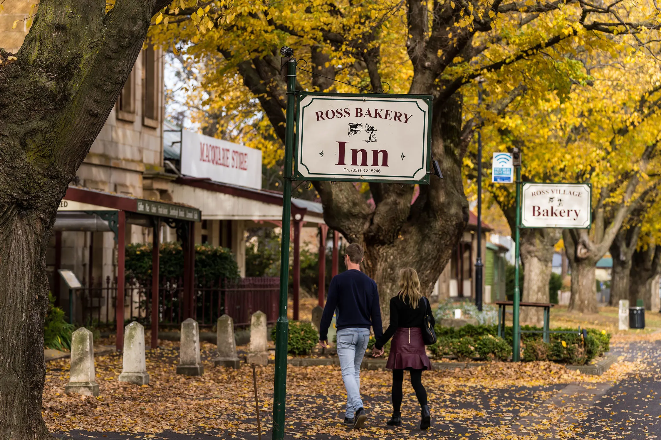 Two people walk along a country town street, with huge yellow-leaved trees surrounding them. Quaint signs hang by the roadside saying 'Ross Bakery Inn' and 'Ross Village Bakery'.