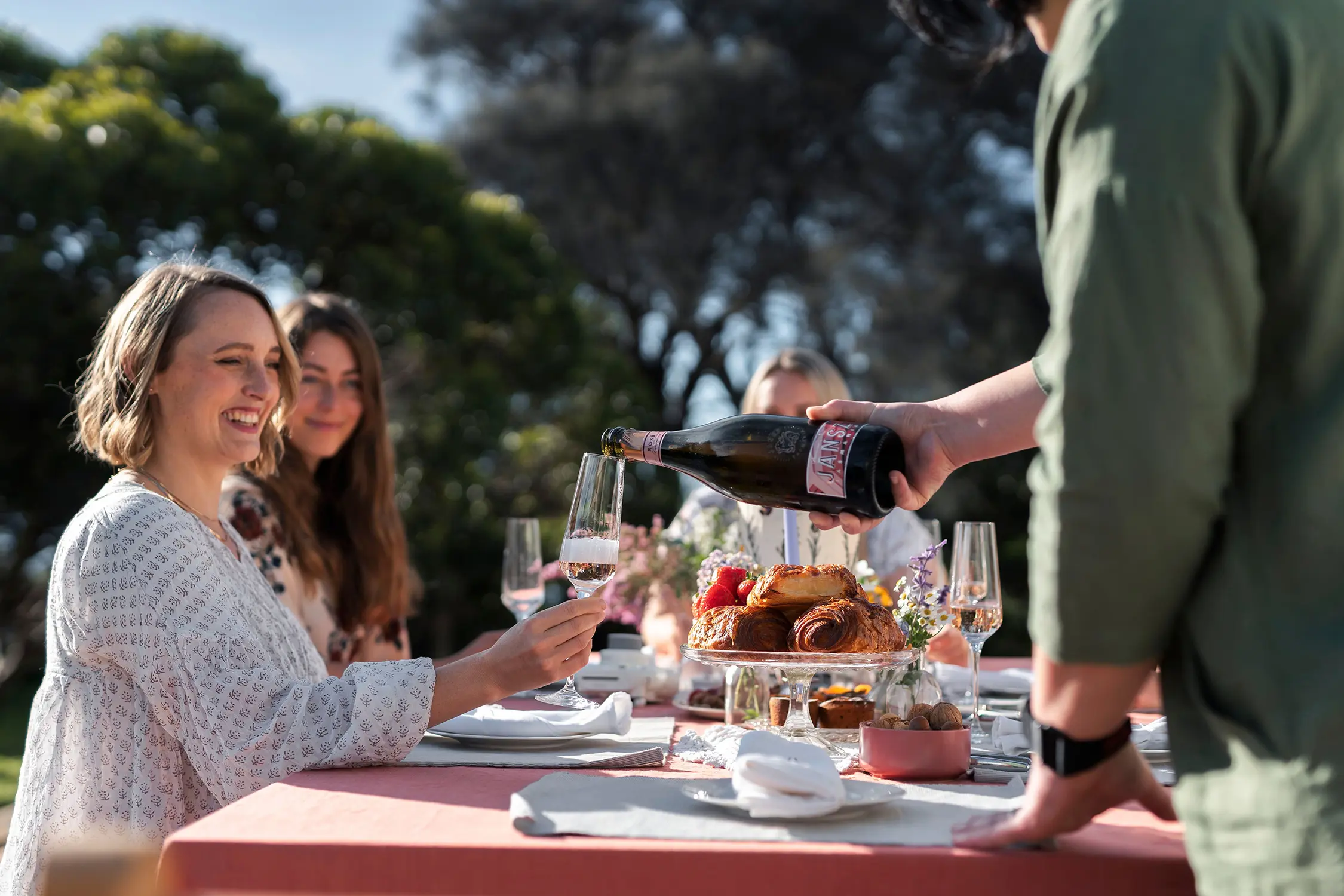 A person leans over a picnic table, set with elegant plates, napkins and floral arrangements, to pour sparkling wine into glasses. 