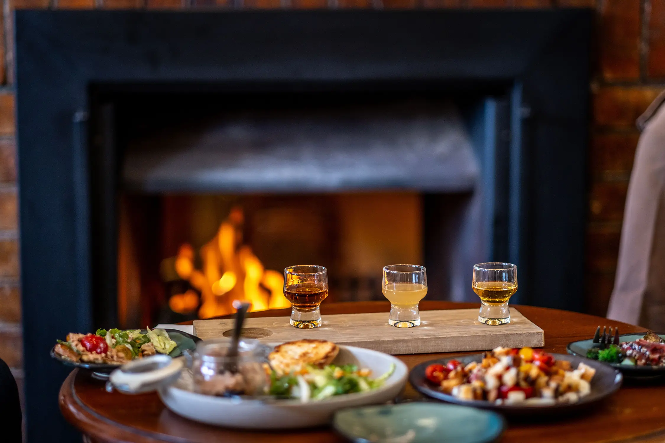 A whisky tasting flight and plates of delicious food sit on a table with a roaring fireplace in the background.
