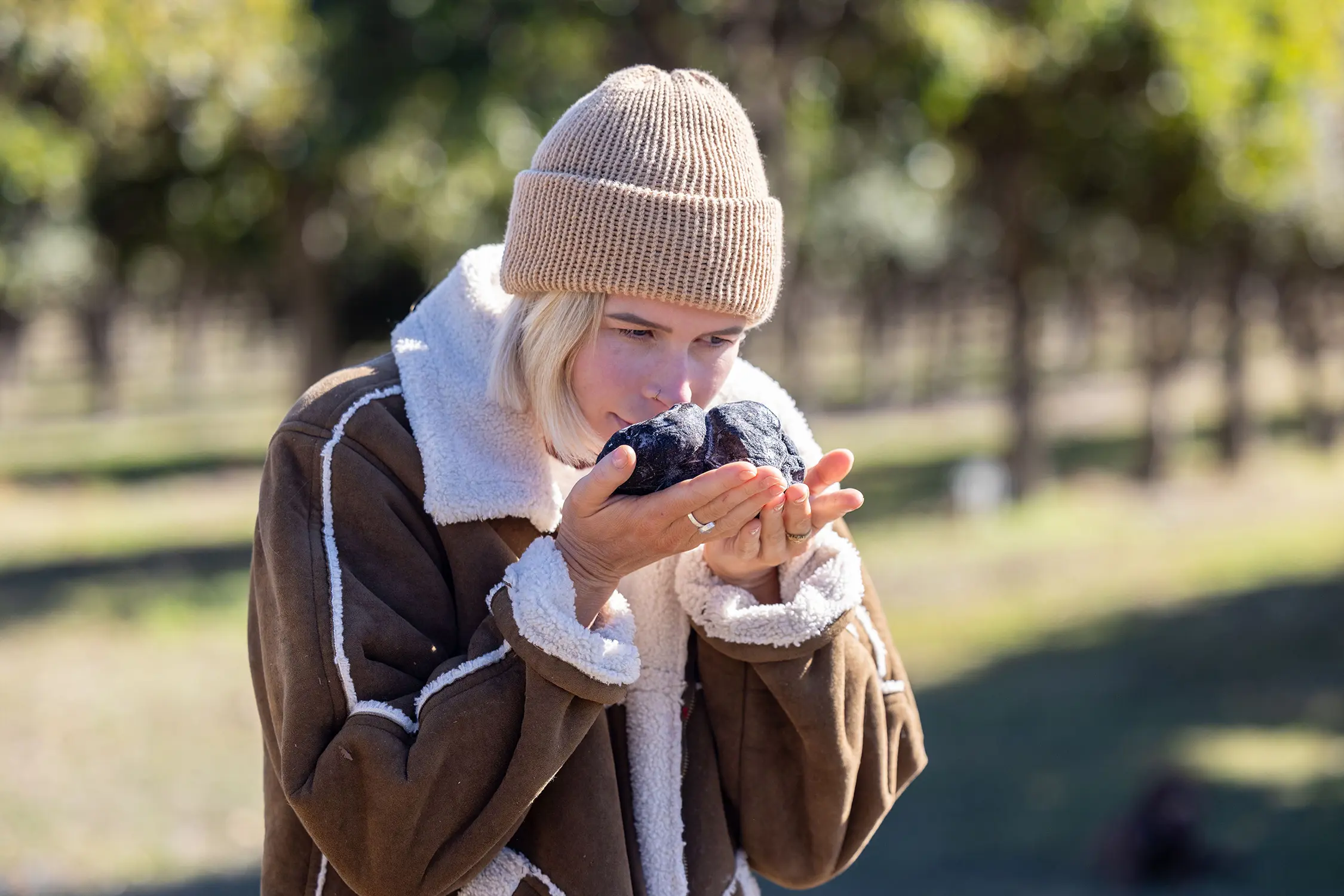 Outside in an orchard, a woman in a jacket and beanie uses both hands to hold a huge truffle up to her face, smelling deeply.