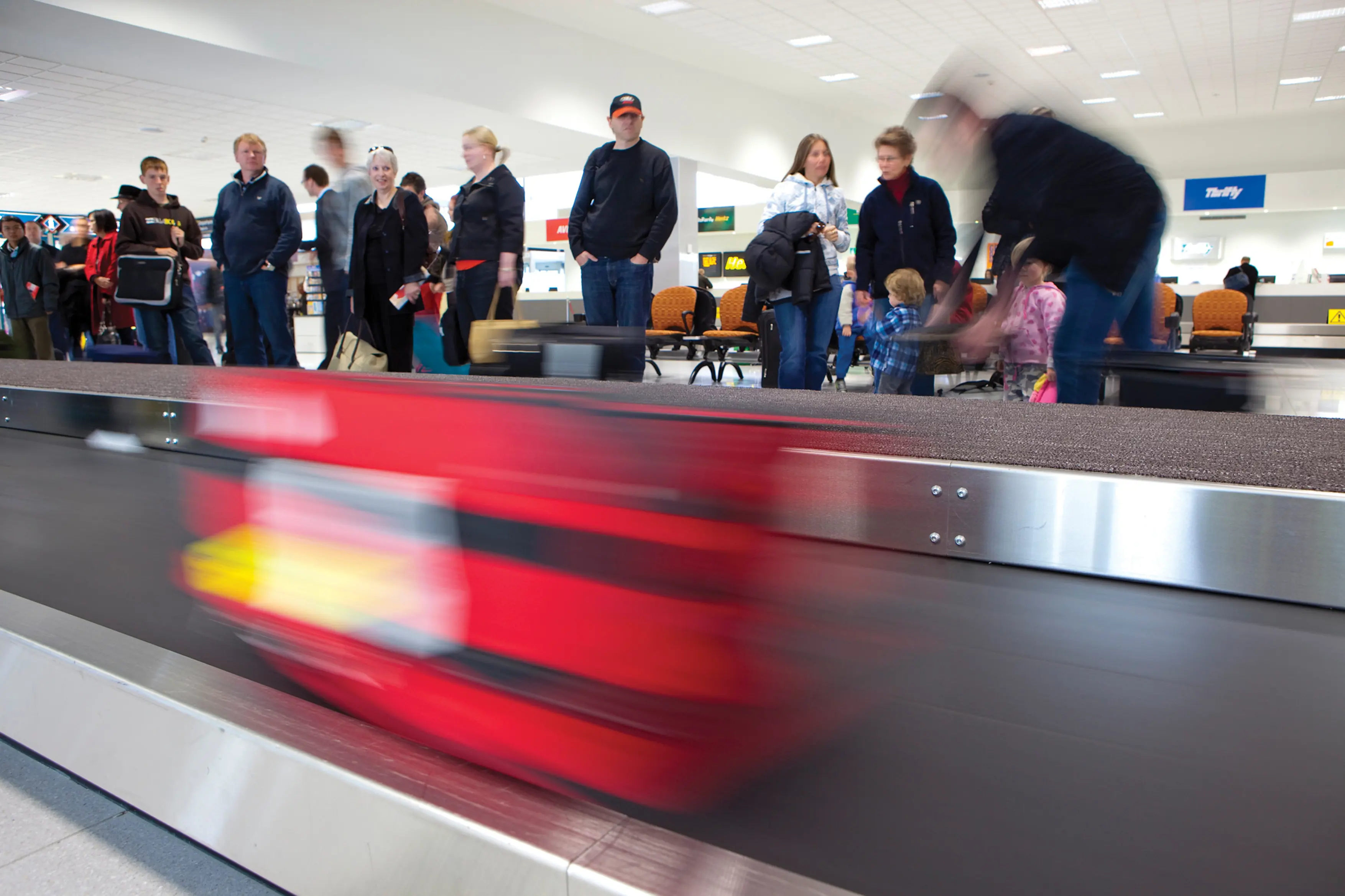 Travellers collect their luggage at Launceston Airport. A bright red suitcase is blurred on the luggage transit in the foreground.
