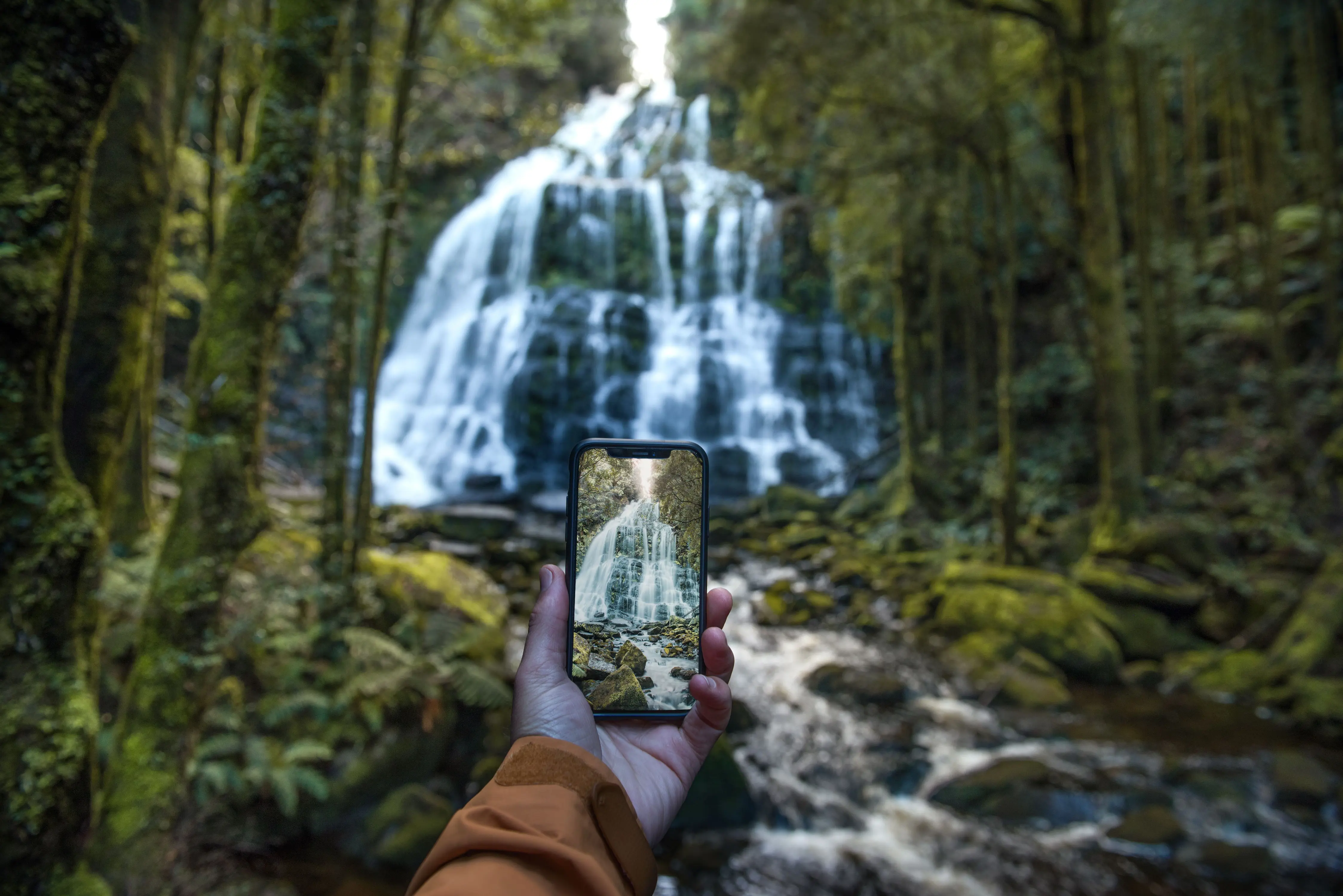 A person's hand holds a mobile phone taking a photo of a multi-tier waterfall framed by green rainforest. The waterfall cascades into a creek lined with mossy rocks.